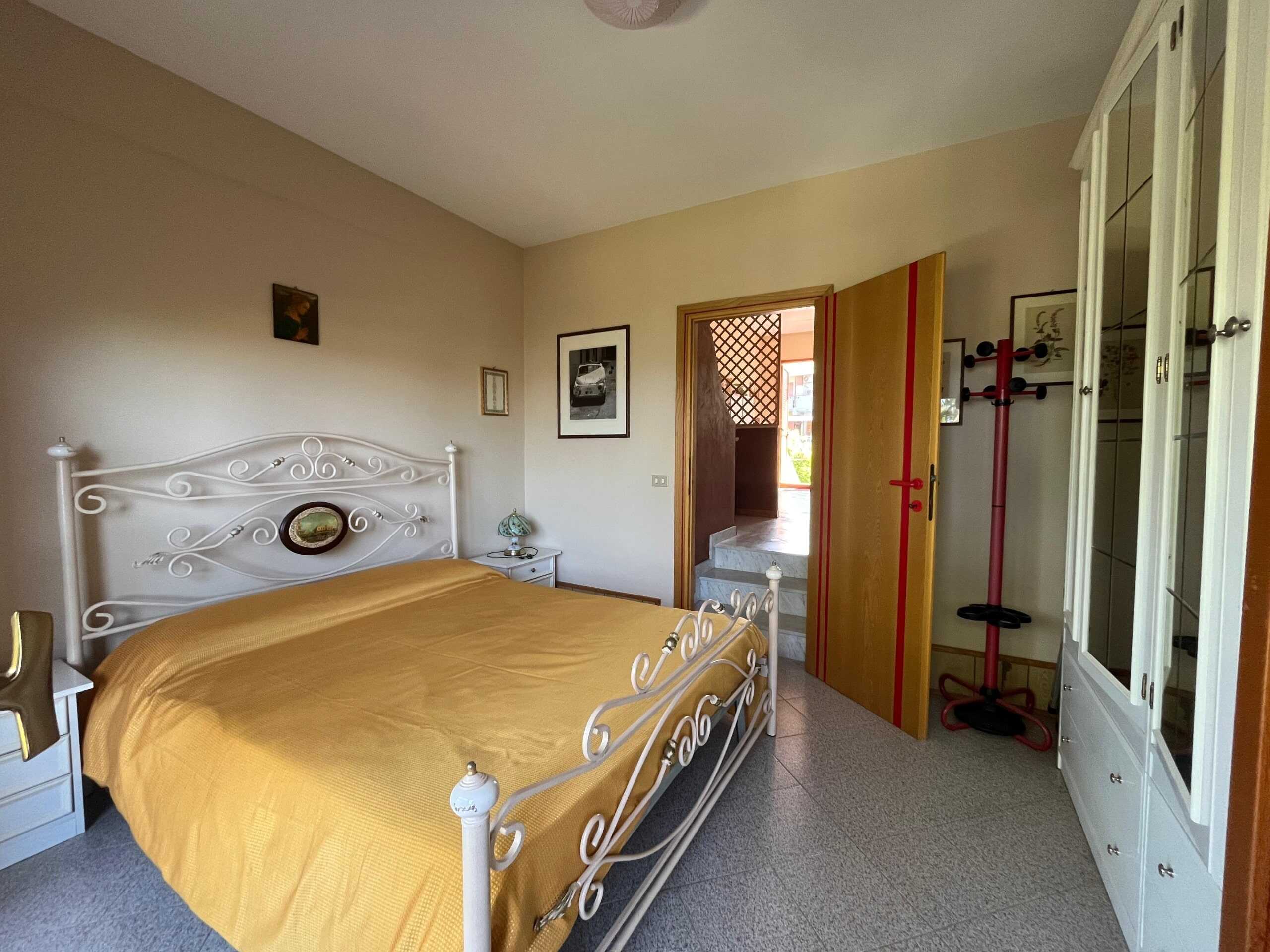 Letto matrimoniale con testiera in ferro battuto bianco e coperta gialla, comodini con lampade, armadio a muro e ingresso al balcone. Arredamento semplice e luminoso in camera da letto, perfetta per un'abitazione confortevole.