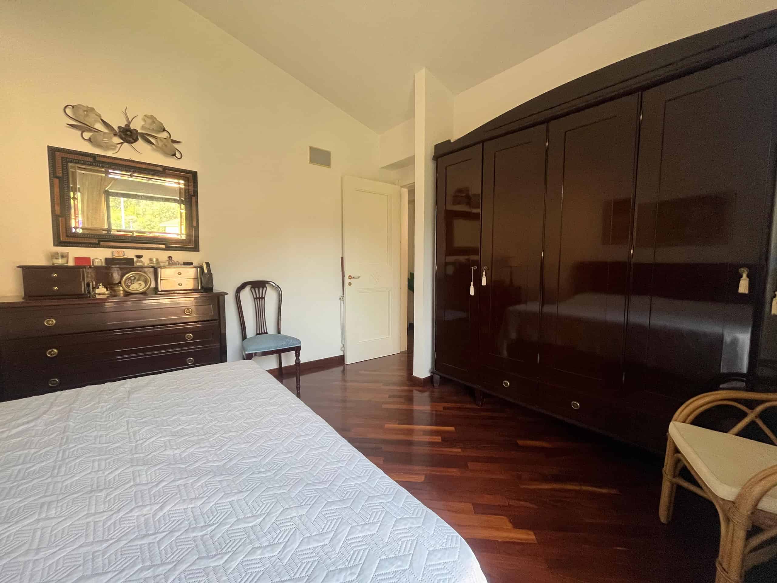 Luci e arredi classici in camera da letto con armadio in legno scuro, letto con coperta bianca, e dettagli decorativi eleganti, ideale per investimenti immobiliari.