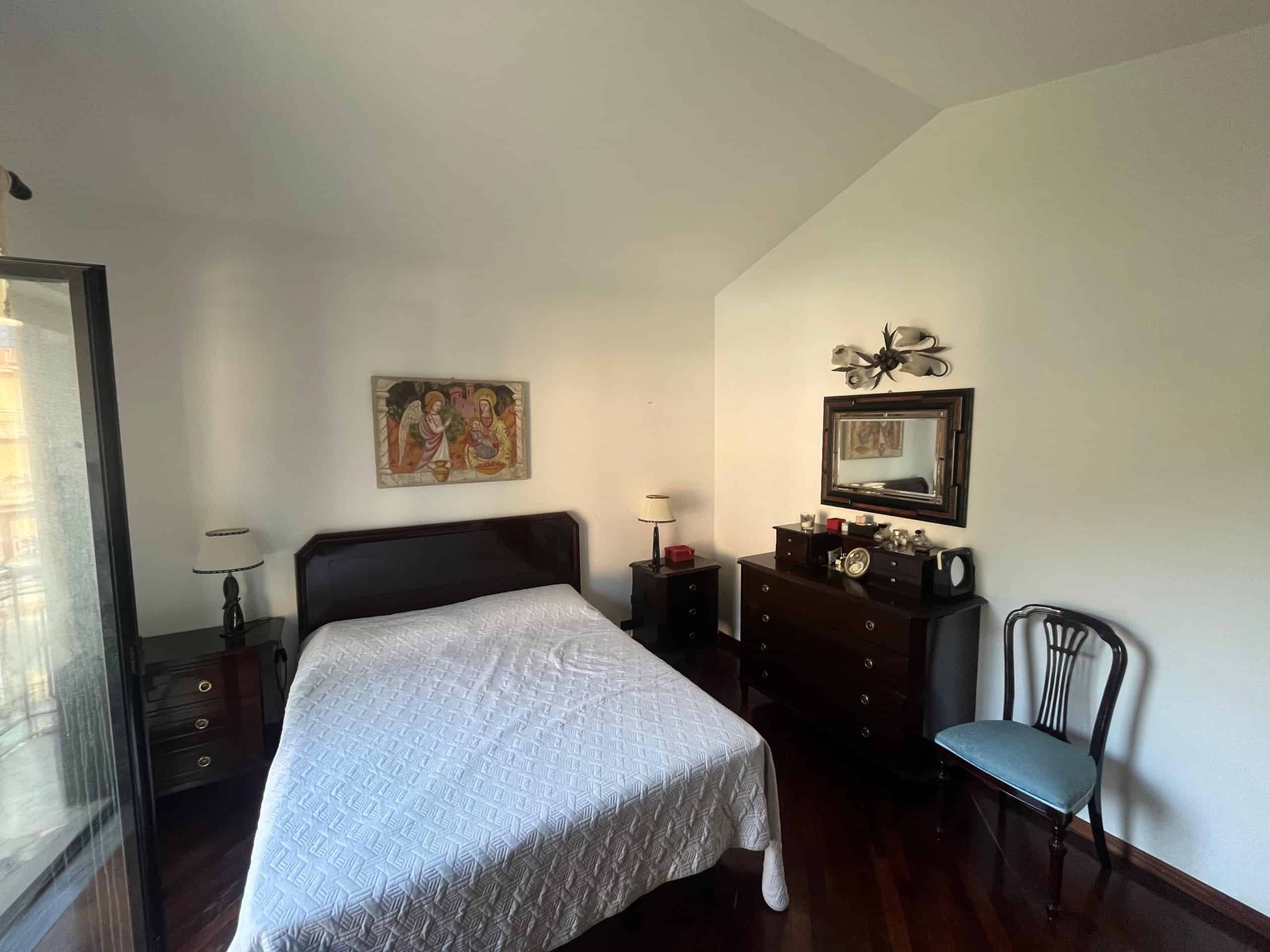 Letto matrimoniale elegante con testiera in legno scuro, arredamento classico, specchio grande, mobile Multiuso, e sedia in stile vintage, ideale per camere da letto di charme.