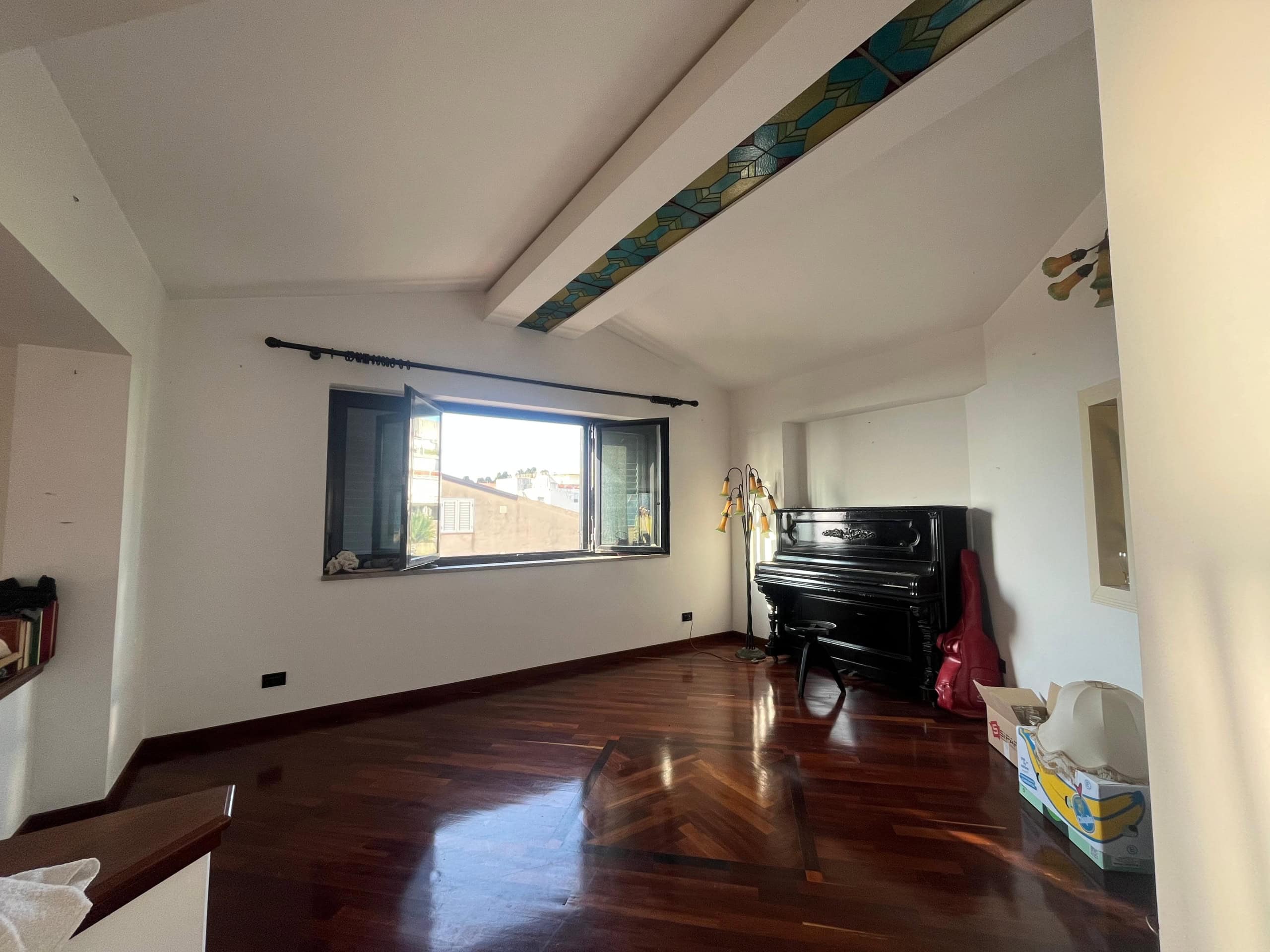 Elegante soggiorno con grande finestra e piano in legno lucido, presente un pianoforte nero, lampada moderna e scatole in stile decorativo, ideale per appartamenti di lusso e riqualificazioni di interni.