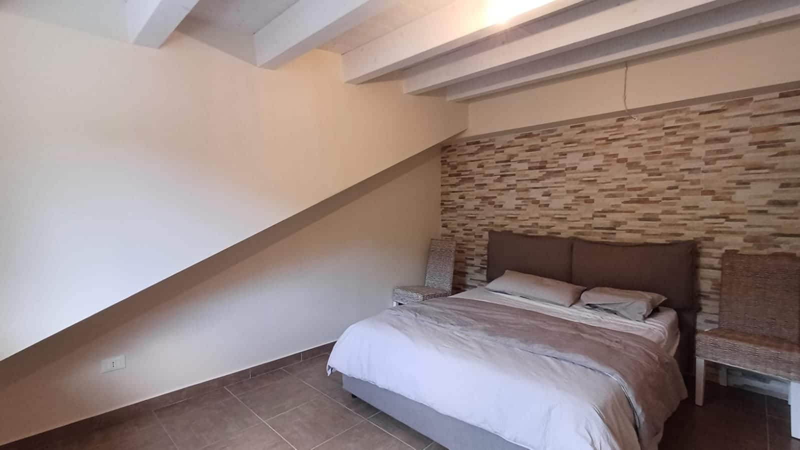 Camera da letto moderna con parete in pietra, letto matrimoniale e cuscini, ambiente accogliente e luminoso, soluzione di arredamento elegante e funzionale, ideale per case di charme.