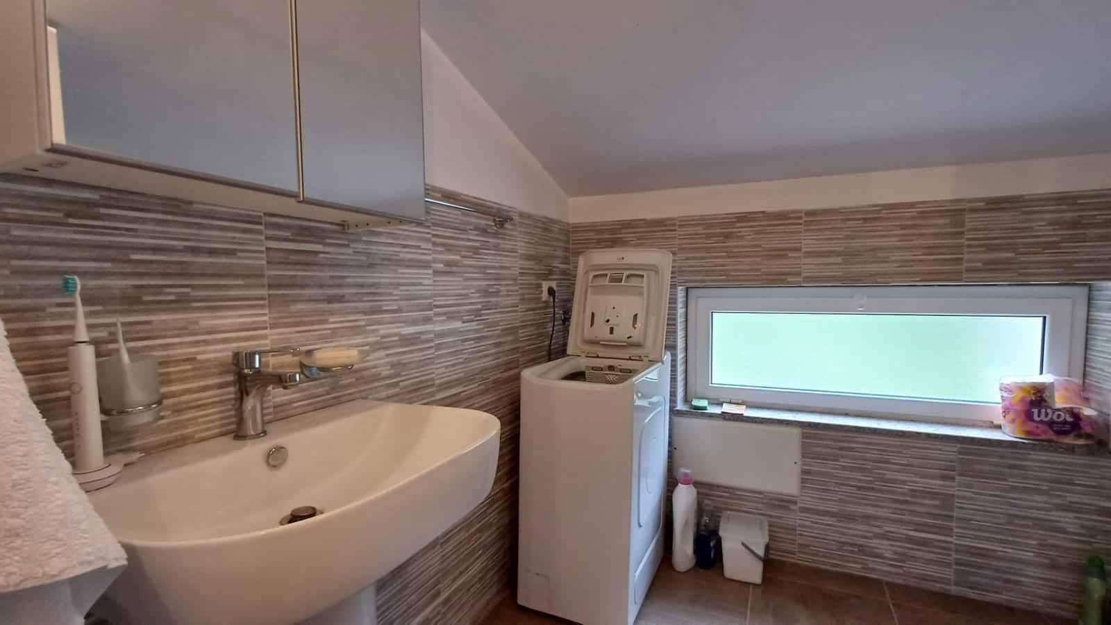 Lavabo moderno in bagno con pareti rivestite di piastrelle in stile moderno, piccolo elettrodomestico di lavanderia vicino a finestra alta, atmosfera luminosa e funzionale.