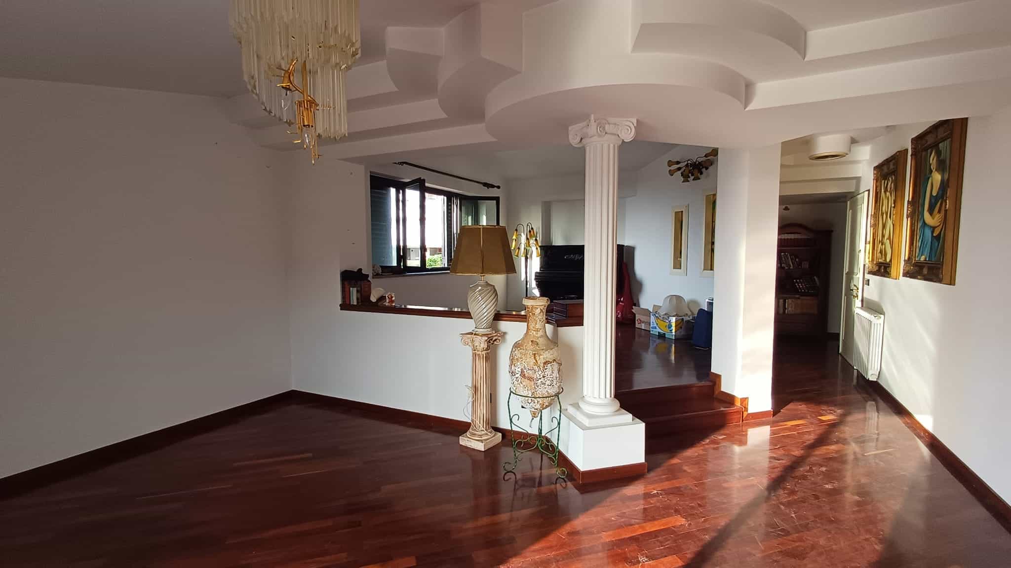 Elegante soggiorno con pareti bianche, pavimento in parquet scuro e dettagli decorativi classici, tra cui colonne e lampadario, ideale per case di alta qualità e arredamento di prestigio.
