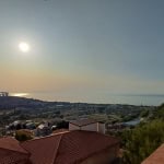 Vista panoramica dal balcone con tetto in tegole rosse, mare e cielo al tramonto, paesaggio collinare e infrastrutture cittadine, ambiente tranquillo e suggestivo in zona residenziale vicino al mare.