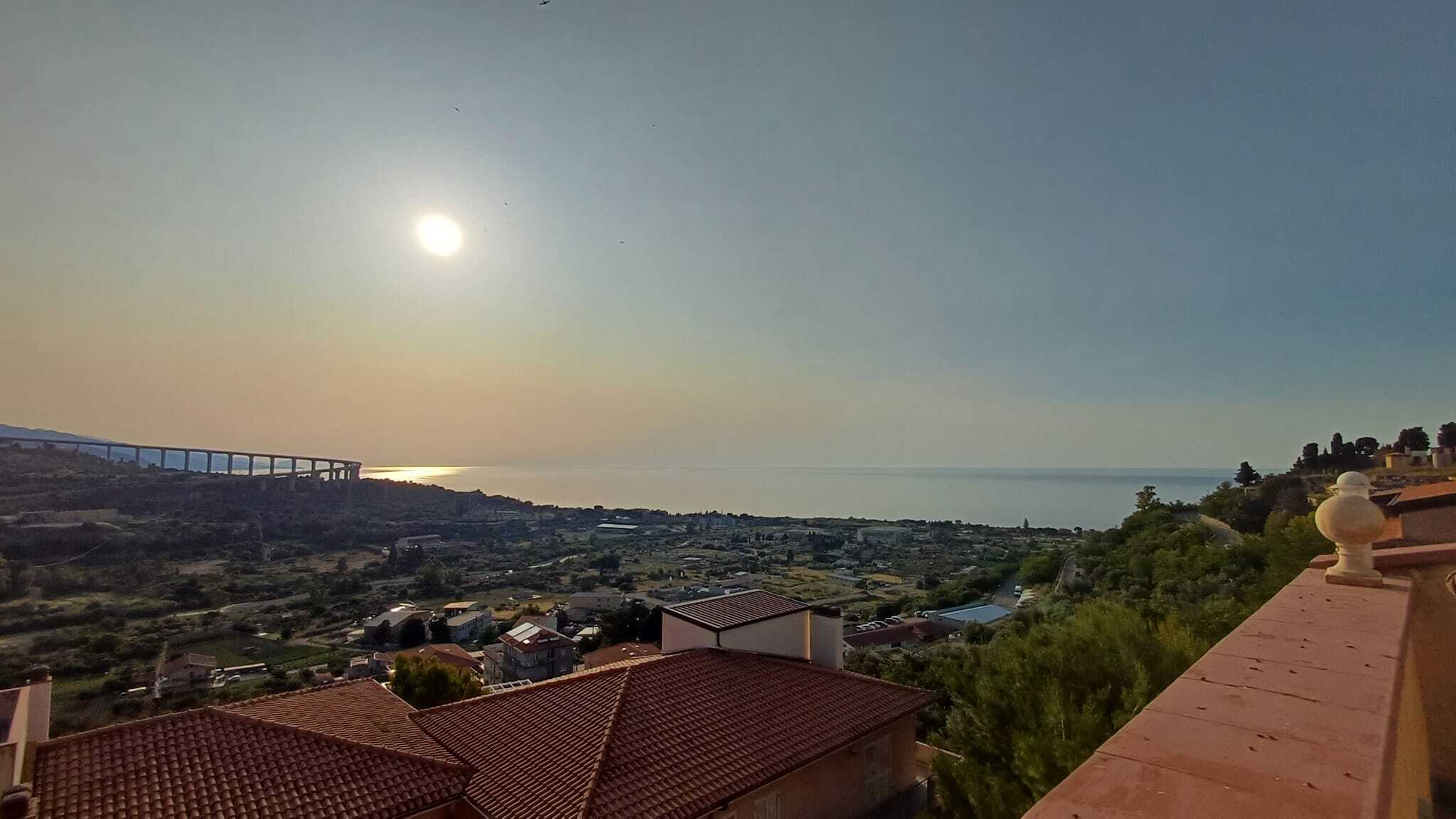 Vista panoramica dal balcone con tetto in tegole rosse, mare e cielo al tramonto, paesaggio collinare e infrastrutture cittadine, ambiente tranquillo e suggestivo in zona residenziale vicino al mare.