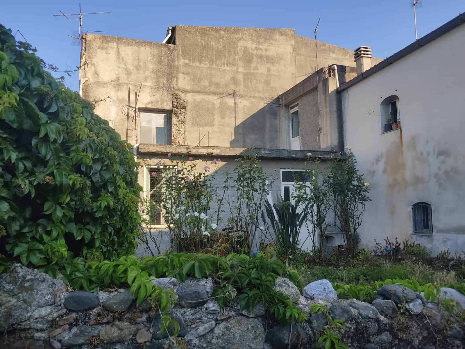 Vecchio edificio residenziale con giardino e vegetazione, facciata in pietra e muratura, in stile rustico, ideale per ristrutturazione, situato in zona tranquilla, con elementi storici e natura.