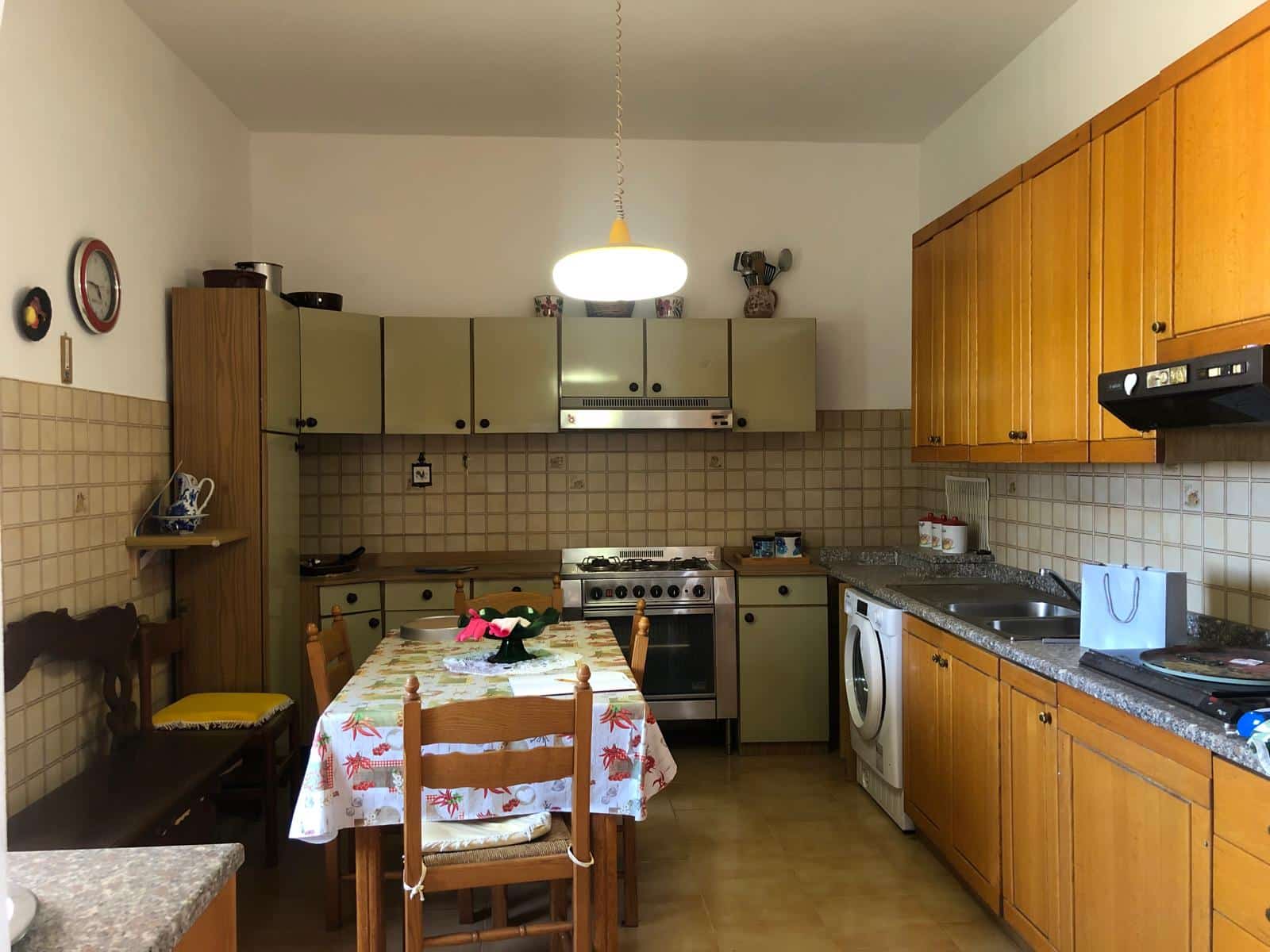Cucina abitabile con mobili in legno e pareti rivestite di piastrelle beige, tavolo con tovaglia decorata, elettrodomestici e illuminazione centrale, atmosfera calda e accogliente.