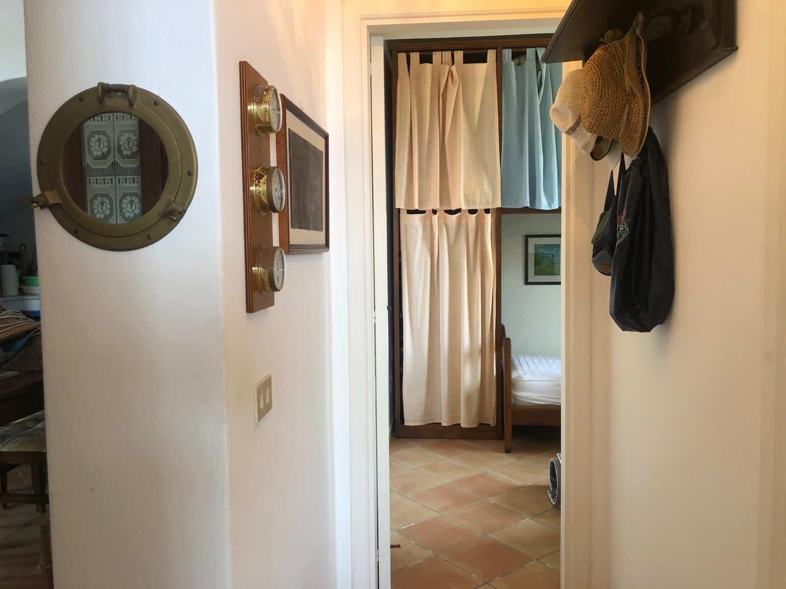 Ampia immagine di un ingresso o corridoio di casa con tende che nascondono una stanza, decorazioni stile marittimo, cappelli e borse appoggiate, ambiente luminoso e accogliente.