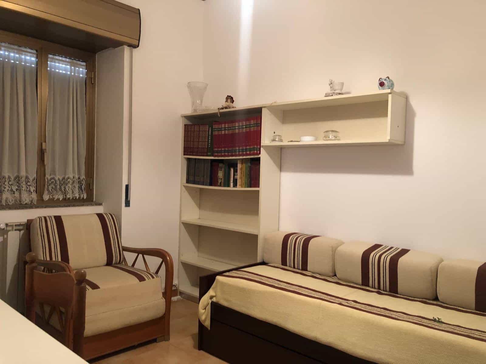 Comforte soggiorno con divano letto e poltrona vintage, libreria a parete con libri e decorazioni, finestra con tende lace, ambiente accogliente e funzionale.