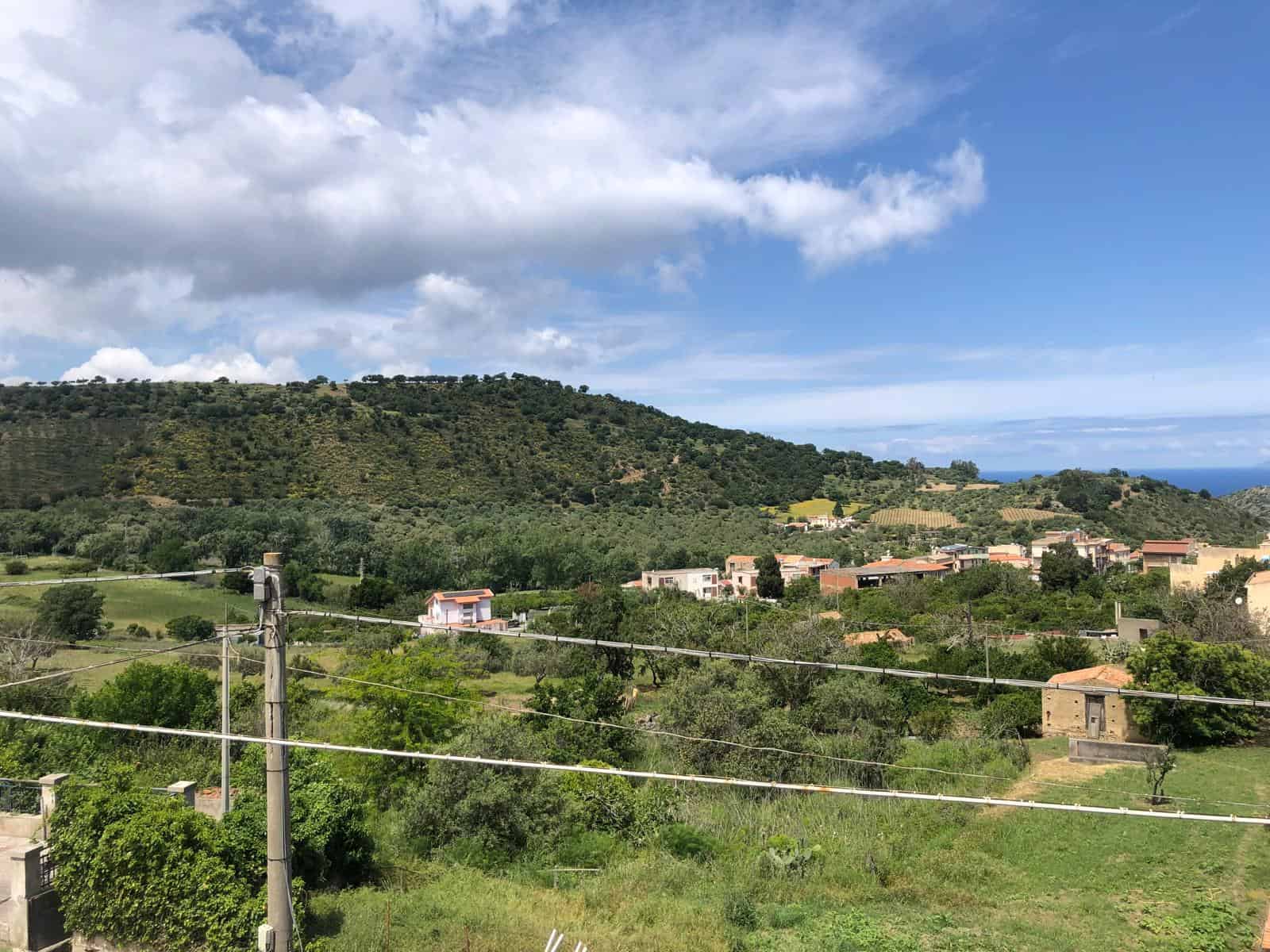 Paesaggio rurale con colline verdi, case e vegetazione, sotto un cielo azzurro con nuvole, ideale per immobili o proprietà di campagna.