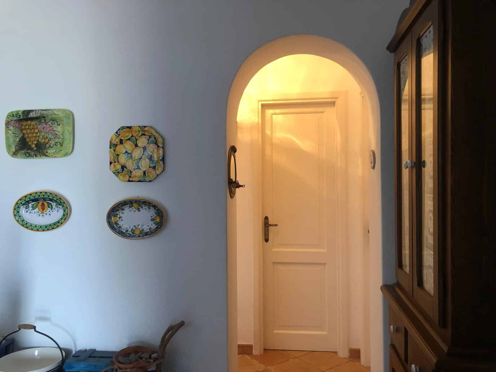 Piatto decorativo in ceramica con motivo di uva e frutta, arredamento rustico in stile mediterraneo, ingresso con porta bianca e arredo classico in legno. Alta qualità, perfetto per ambienti casalinghi accoglienti.