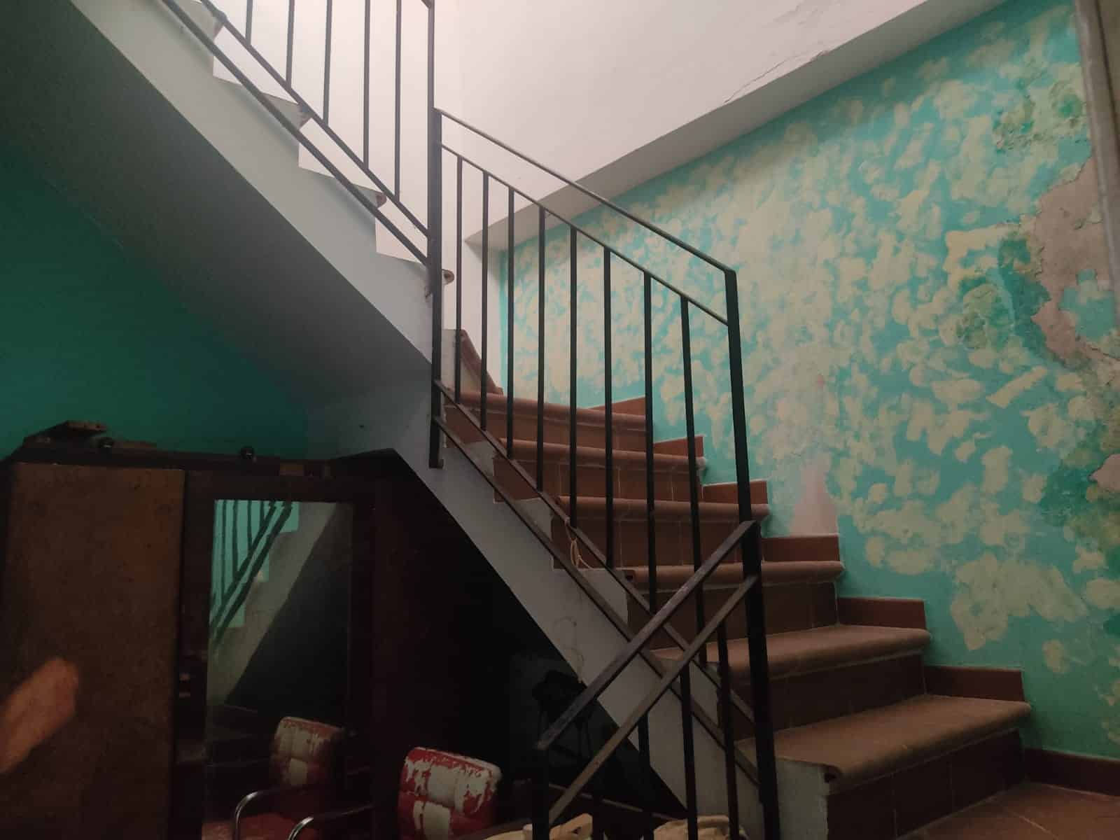 Scala in muratura con ringhiera in metallo, pareti con vernice a spruzzo verde acqua e cemento sfaldato, ambiente interno di una proprietà in vendita o in ristrutturazione.
