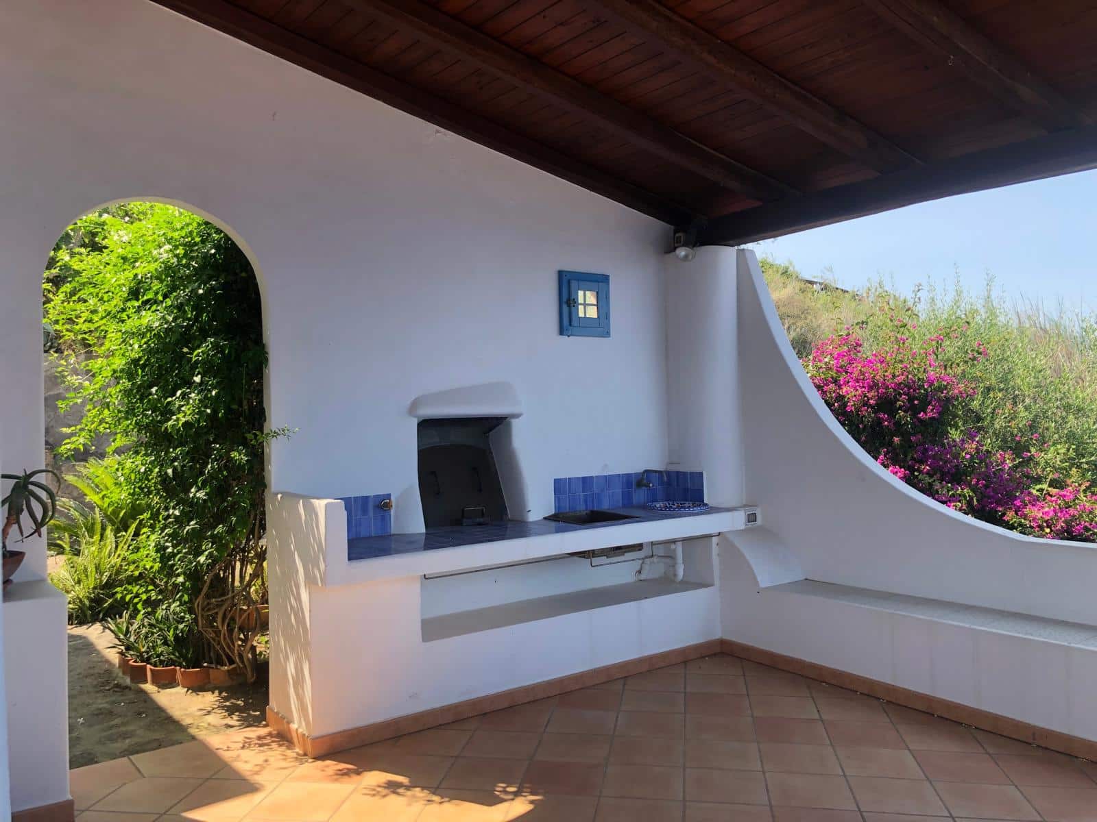 Spazio esterno con barbecue in stile mediterraneo, pareti bianche e dettagli blu, vista su fiori colorati e natura, perfetto per cene all'aperto e relax in un ambiente accogliente e autentico.