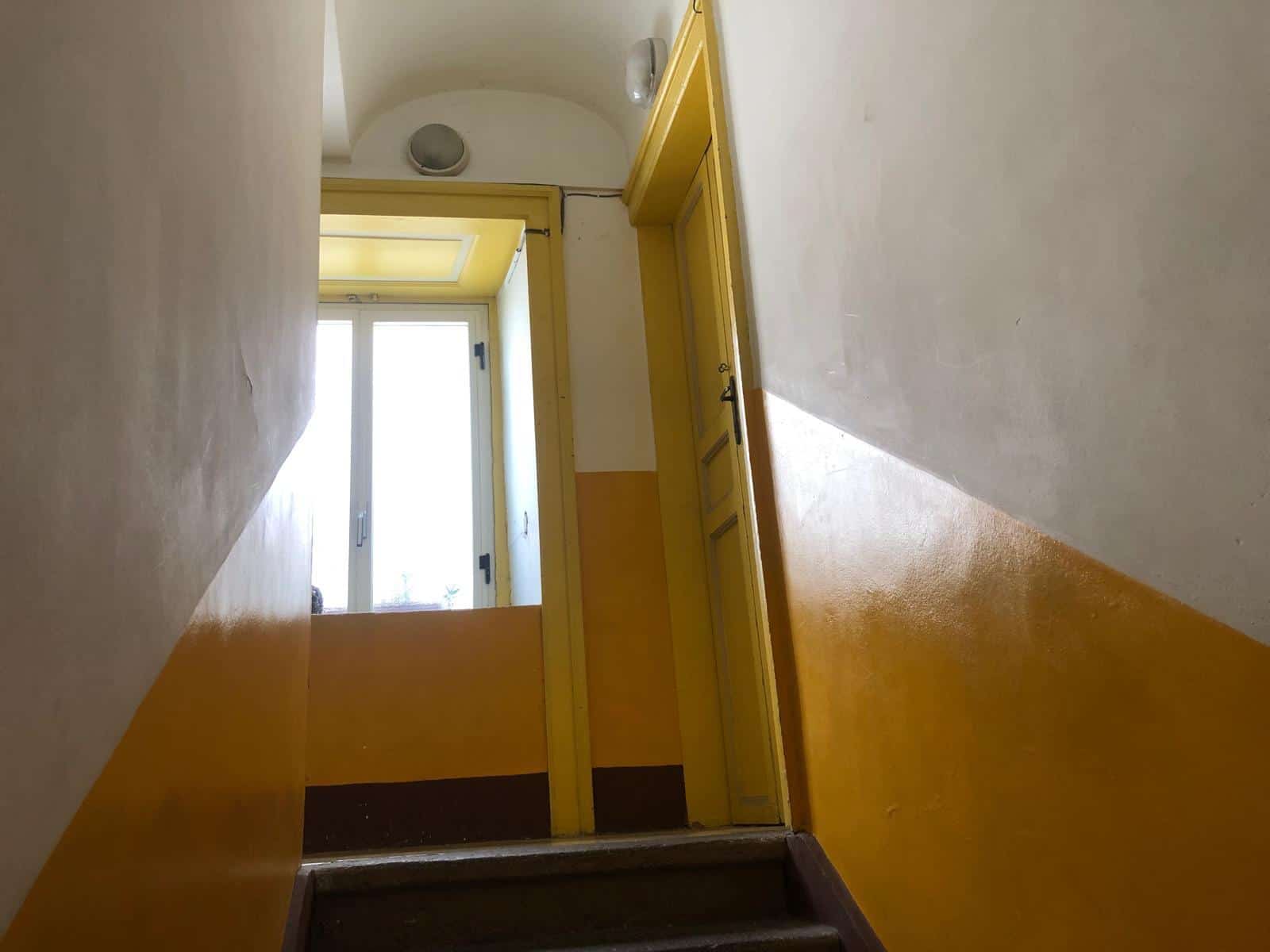 Ingresso di un appartamento con porta gialla, pareti bianche e soffitto arcuato, illuminato dalla luce naturale della finestra.