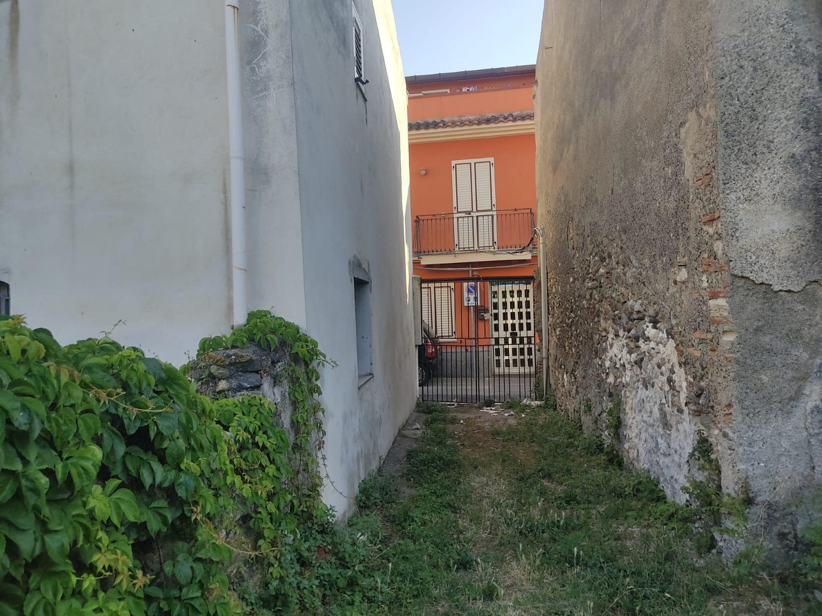 Ingresso di cortile tra muri con vegetazione e sbarra di ferro, enclavato tra edifici residenziali, in un contesto di quartiere urbano con case colorate e strada asfaltata.