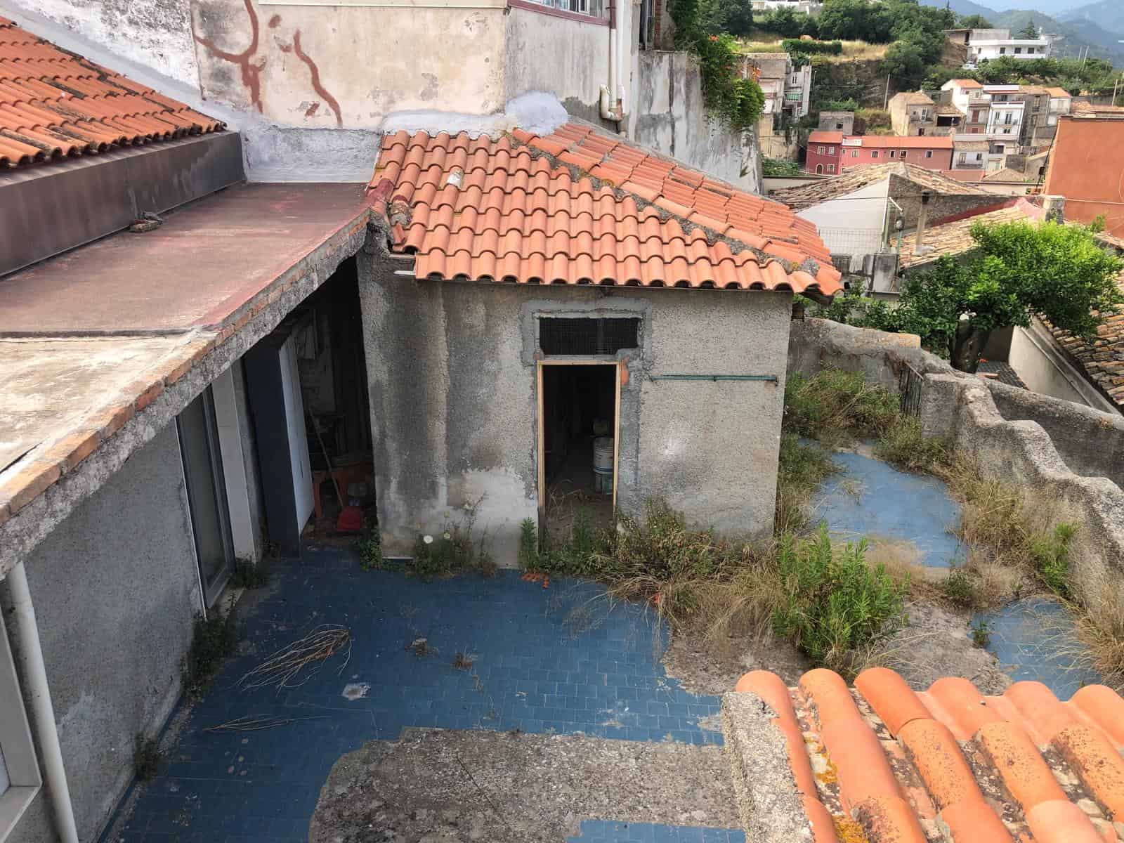Tetto in terracotta e piccolo cortile trasandato con piante cresciute selvaggiamente, vista di un quartiere residenziale su colline, condizione di edificio da ristrutturare, NextCasa immobiliare.