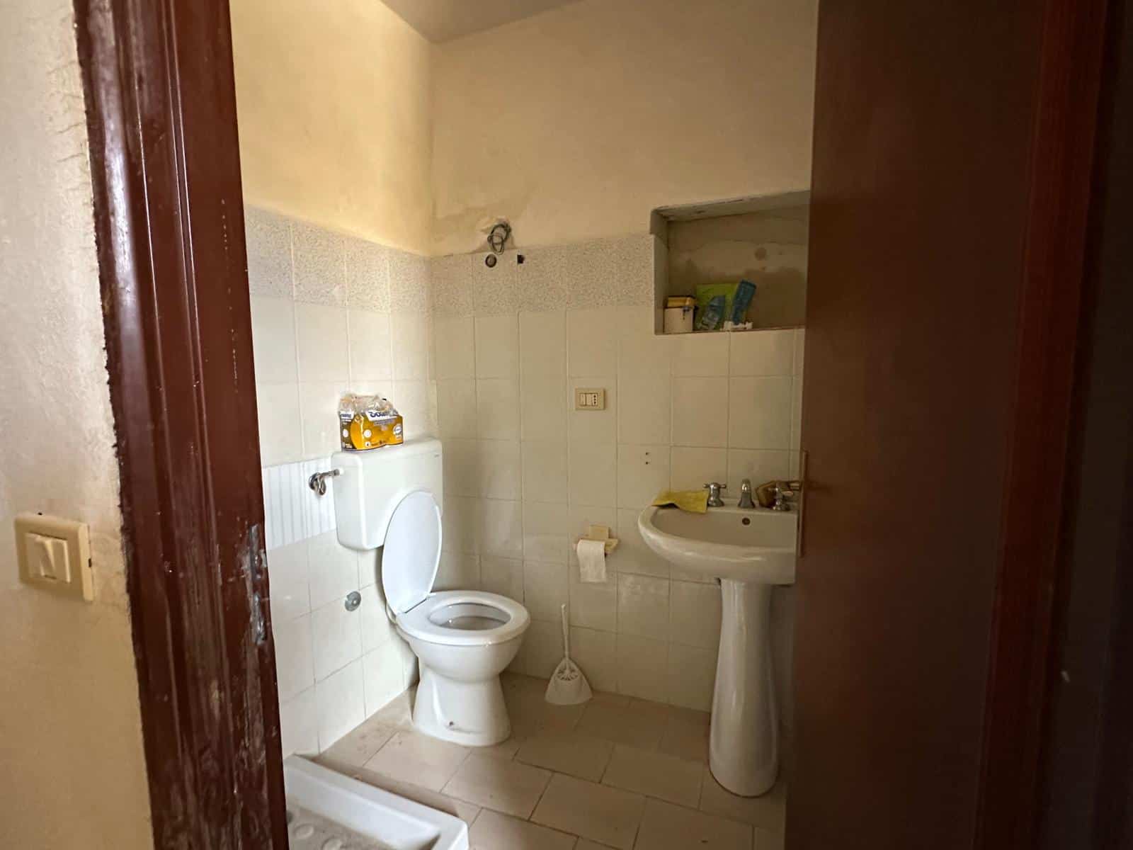 Bagno in fase di ristrutturazione con sanitari nuovi, lavabo, tazza e ripiano con scaffale, pareti rivestite con piastrelle chiare, pavimento in gres, pareti color sabbia e infissi in legno.