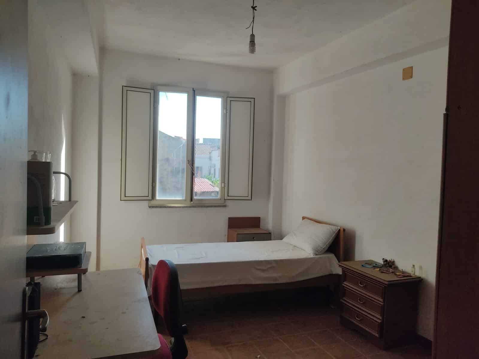 Ampio ambiente abitativo con una camera singola arredata, finestra luminosa, mobili in legno e pareti bianche, ideale per solutioni di affitto o investimento immobiliare.