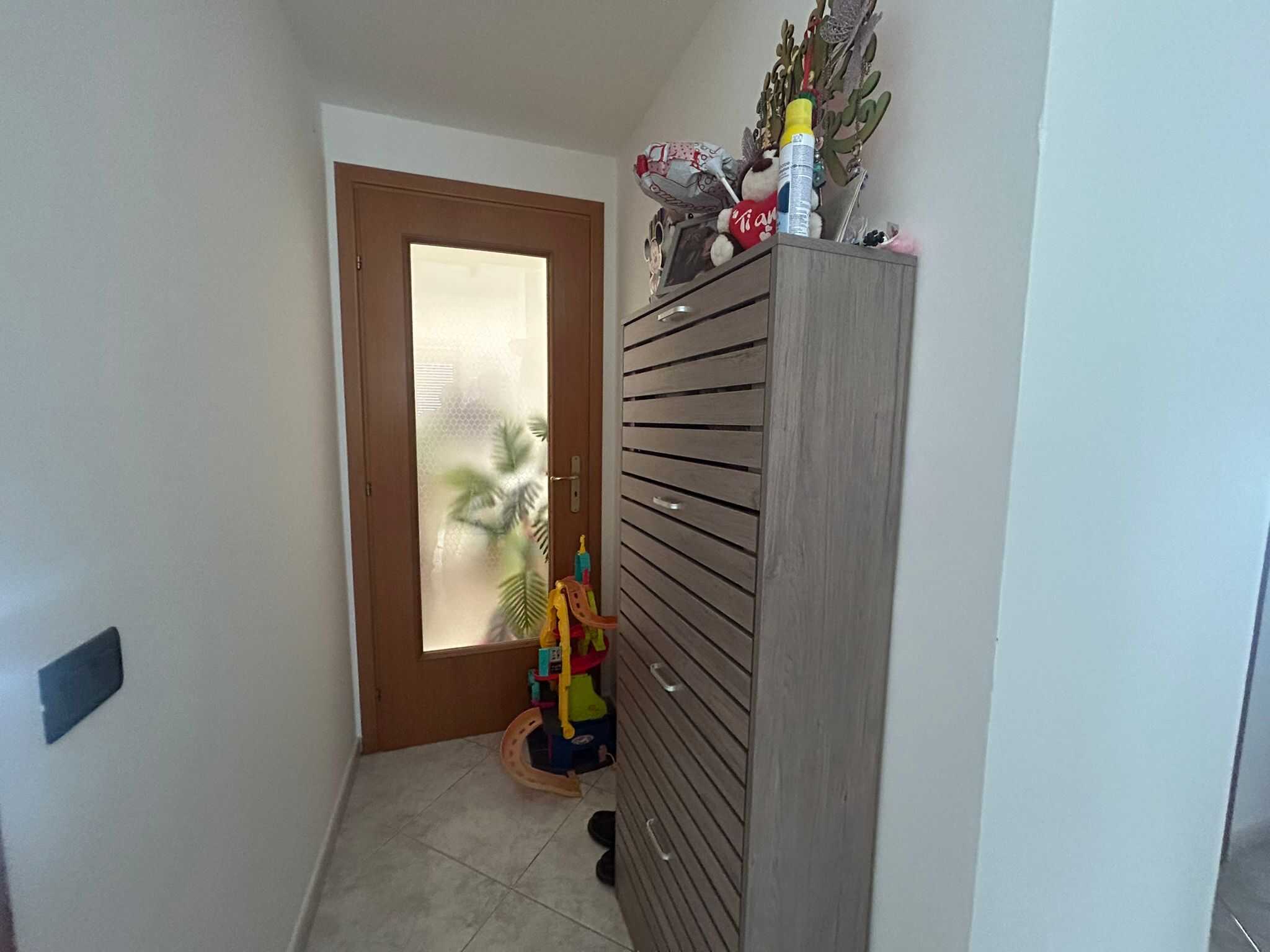 Scarpa da ingresso in legno chiaro con bottega, porta in legno con vetro satinato e decorazioni verdi di piante, arredamento funzionale per l'ingresso di una casa.