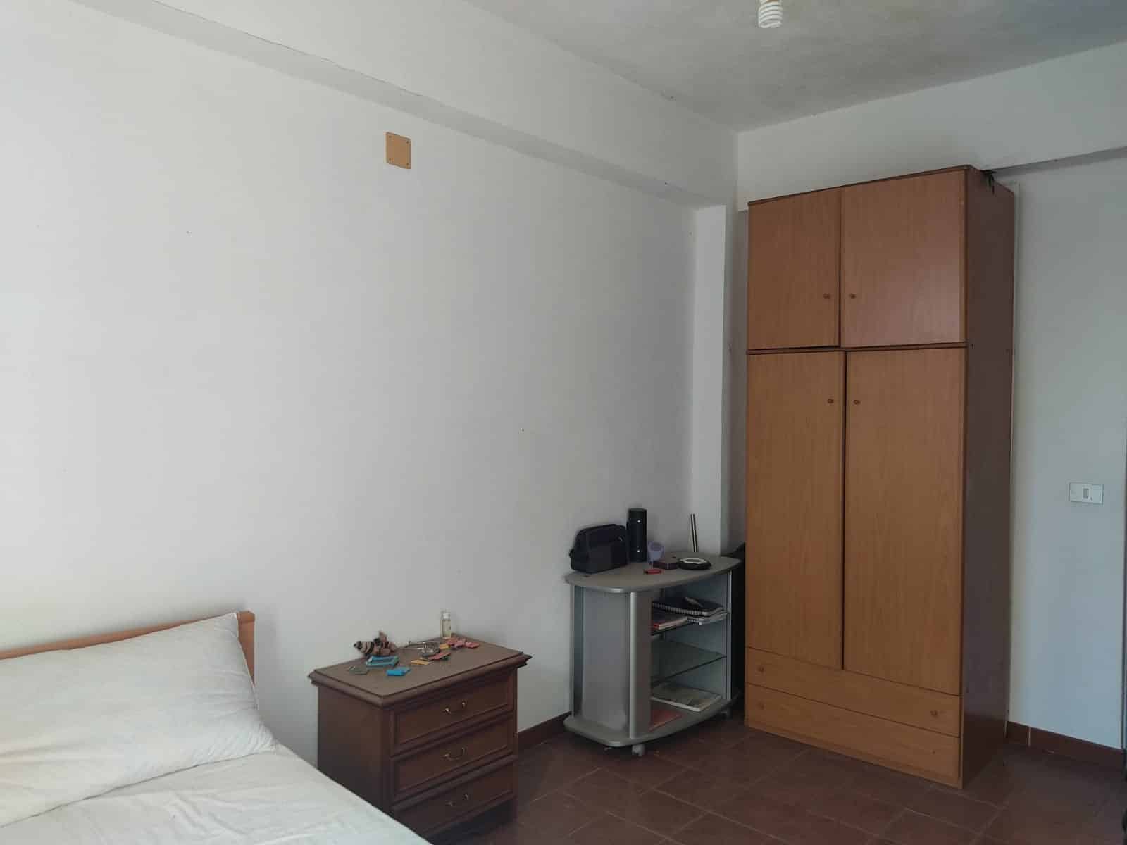 Armadio in legno grande in camera da letto con piccola scrivania e carrozzina accanto, parete bianca e pavimento in piastrelle, stanza semplice e funzionale.