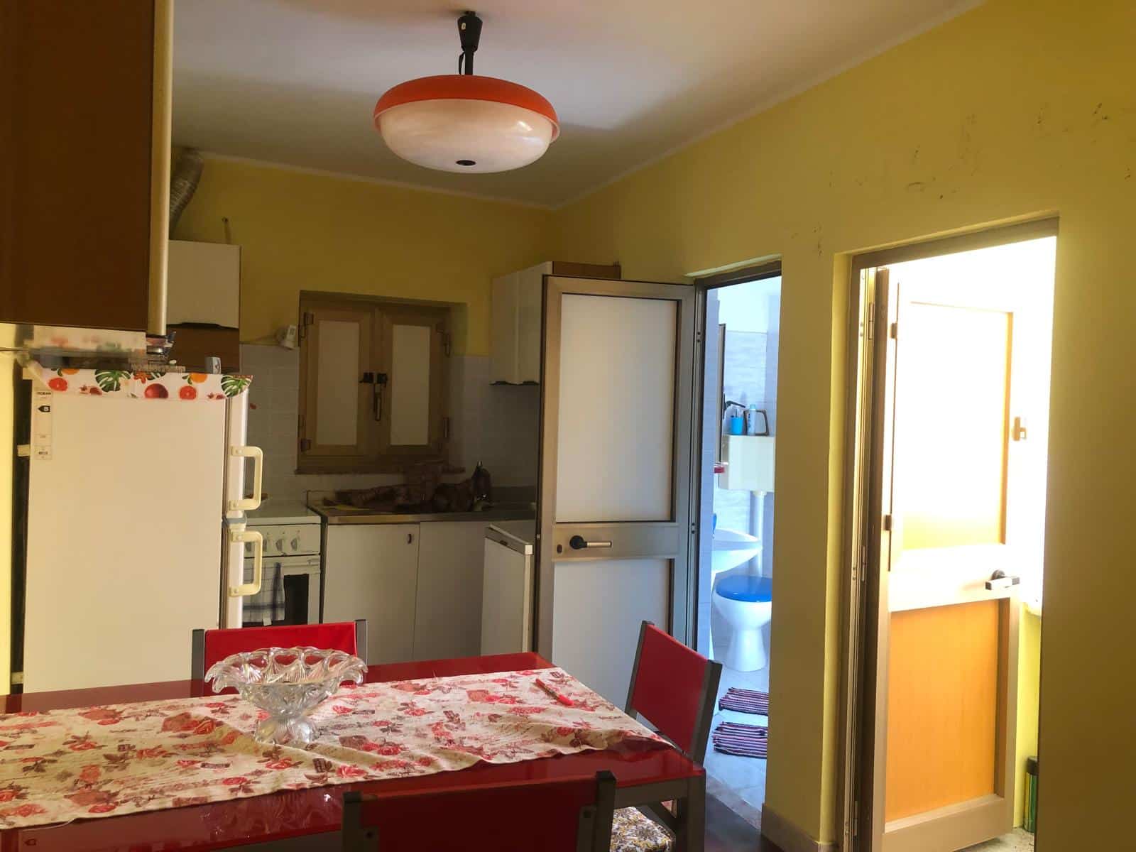 Mini cucina soggiorno con pareti gialle, tavolo rosso con tovaglia floreale, finestra e porta di accesso alla stanza da bagno, ripiani e frigorifero bianco.