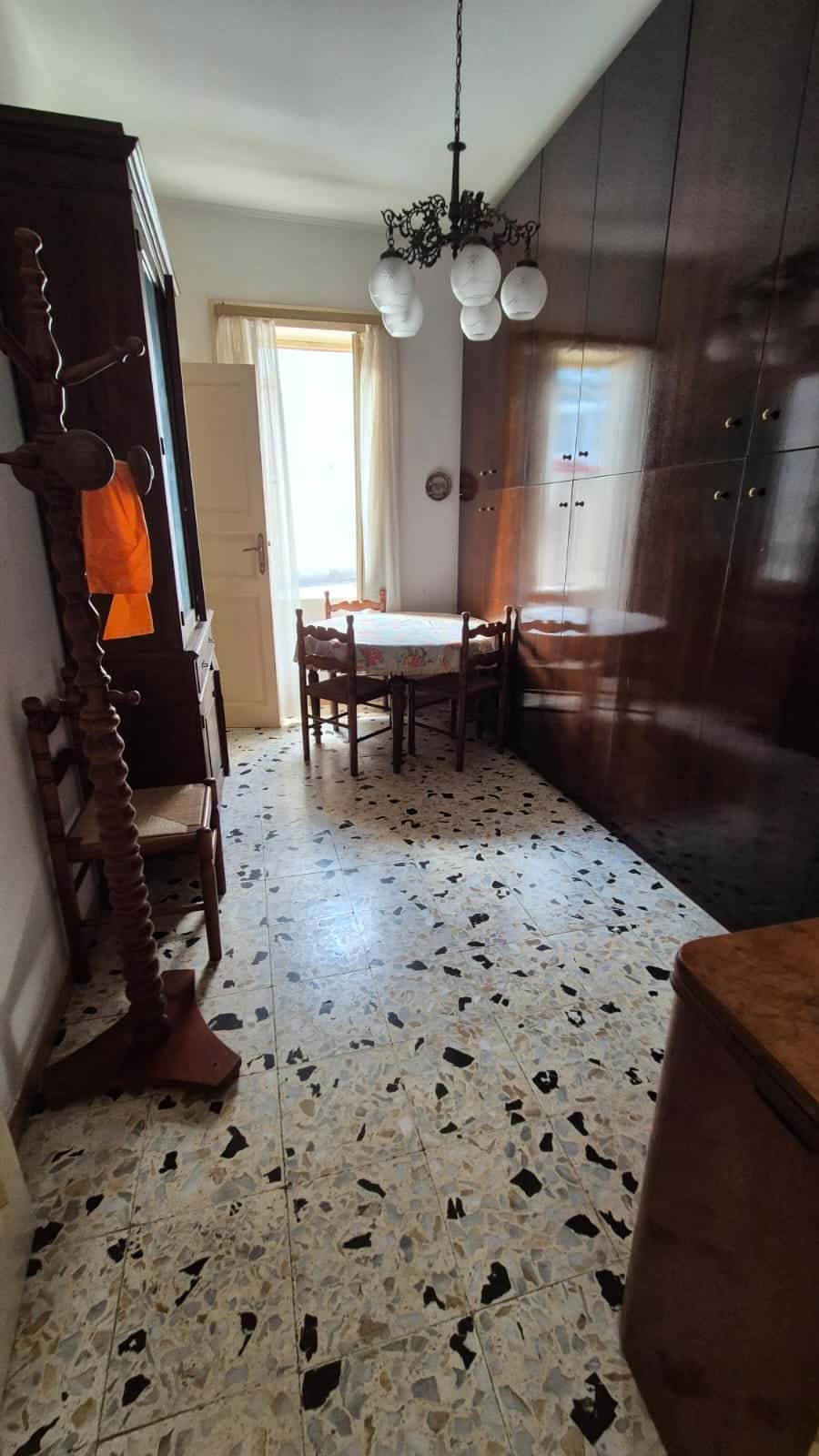 Antico ambiente soggiorno con mobile in legno scuro, tavolo con tovaglia floreale, lampadario vintage a soffitto e pavimento in marmo con mosaico beige e nero.