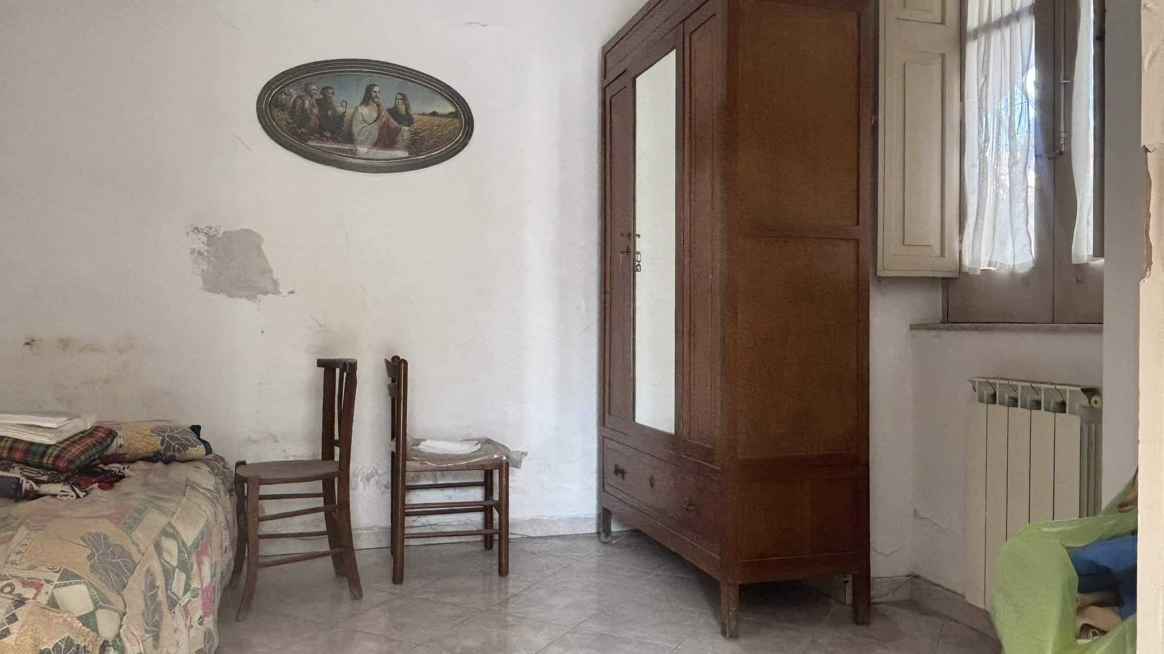 Armadio antico in legno, camera da letto semplice con mobili vintage, pareti rovinate, finestra con tende leggere e radiatore, stile rustico e abitabile.