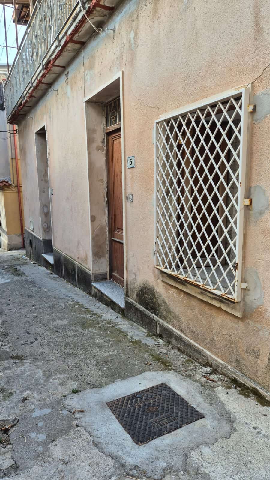 Porte di casa con portoncino in legno e finestra protetta da inferriata bianca, facciata esterna in pietra e cemento, pareti con segni di degrado, piccolo vicolo urbano con pavimentazione sconnessa e tombino in primo piano, immobile da ristrutturare, zona residenziale storica italiana, atmosfera urbana autentica.