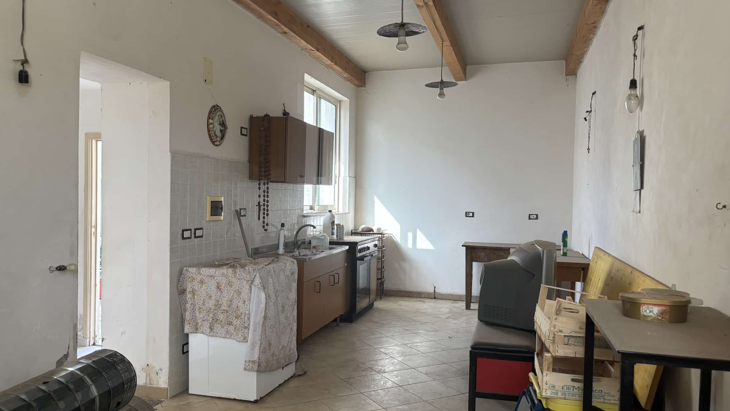 Immagine di una cucina rustica in fase di ristrutturazione, con pareti bianche, soffitto con travi in legno e pavimento in piastrelle. Sono presenti mobili, utensili, e alcuni oggetti di lavoro sparsi, con illuminazione naturale dalla finestra.