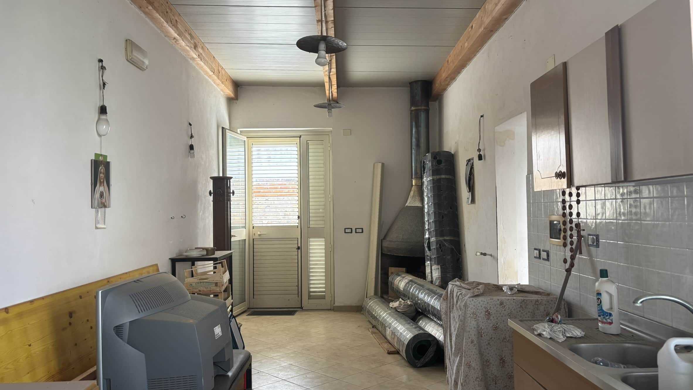 Finestra in cucina con persiana, ambiente da ristrutturare, pareti bianche, tende, ardesia, oggetti di uso quotidiano, forno, lavabo, mobili da cucina, soffitto in legno, un camino, materiali per lavori, illuminazione con lampade sospese.
