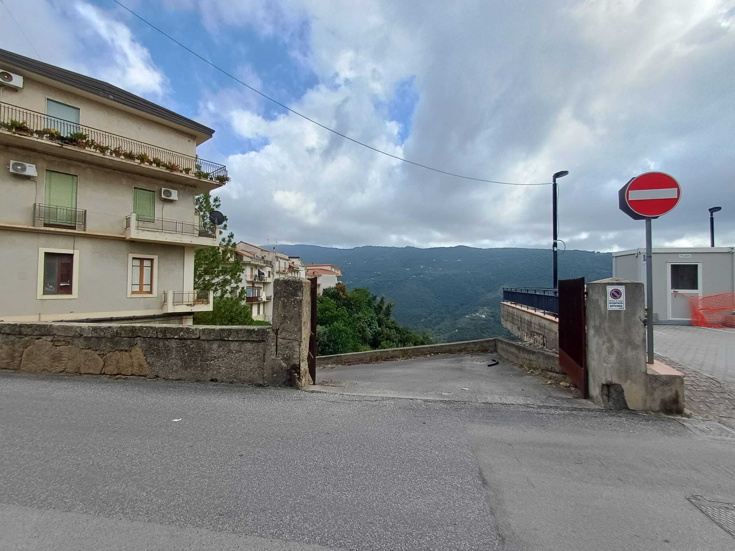 Storico edificio residenziale con vista panoramica sulle colline, strada di accesso in discesa con segnale di divieto di accesso, ambiente urbano con elementi di arredo urbano e paesaggio naturale sullo sfondo.