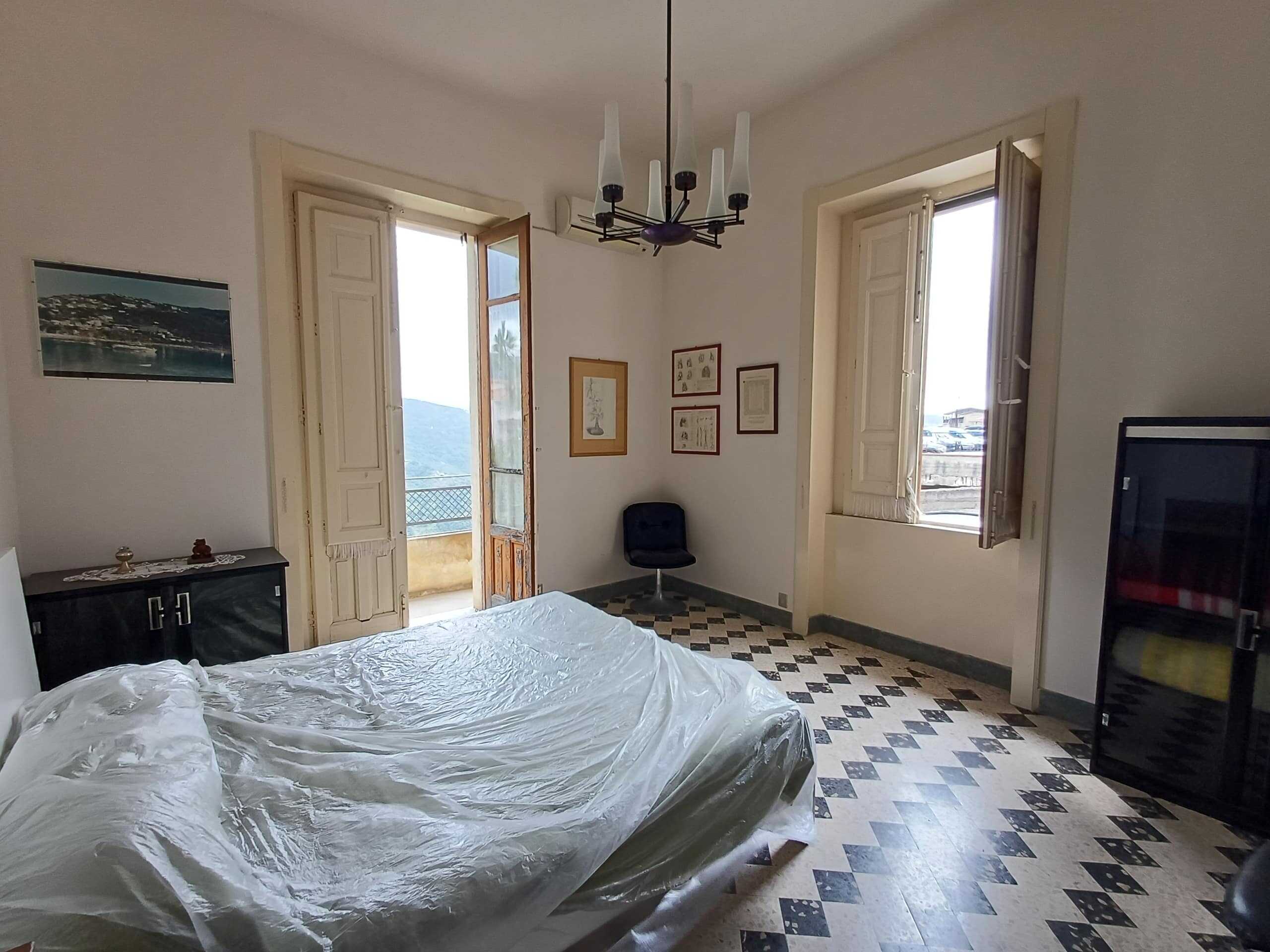 Letto matrimoniale con biancheria in una camera luminosa con pareti bianche e grandi finestre che offrono una vista sul mare e sulle colline circostanti, arredamento semplice ed elegante.