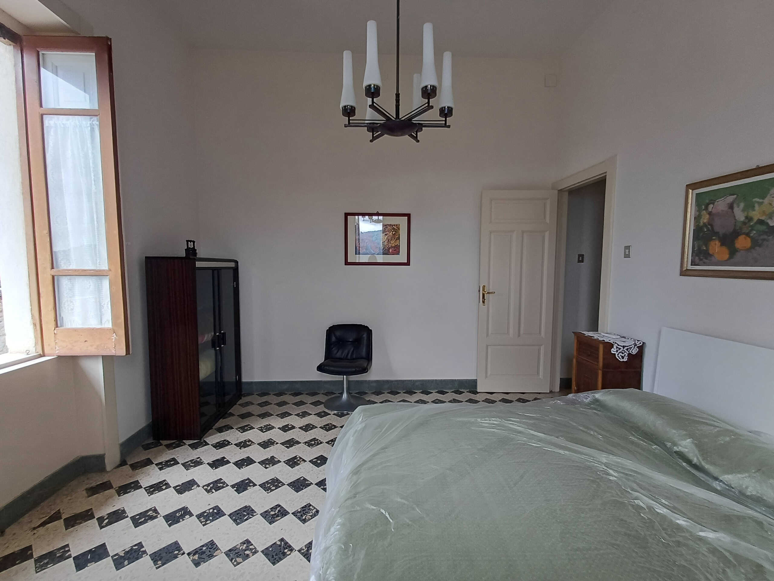 Spazio camera da letto luminoso con pareti bianche, grande finestra, biancheria da letto verde e arredi classici. Interno rustico elegante con dettagli in legno e pavimento in piastrelle neri e bianche.
