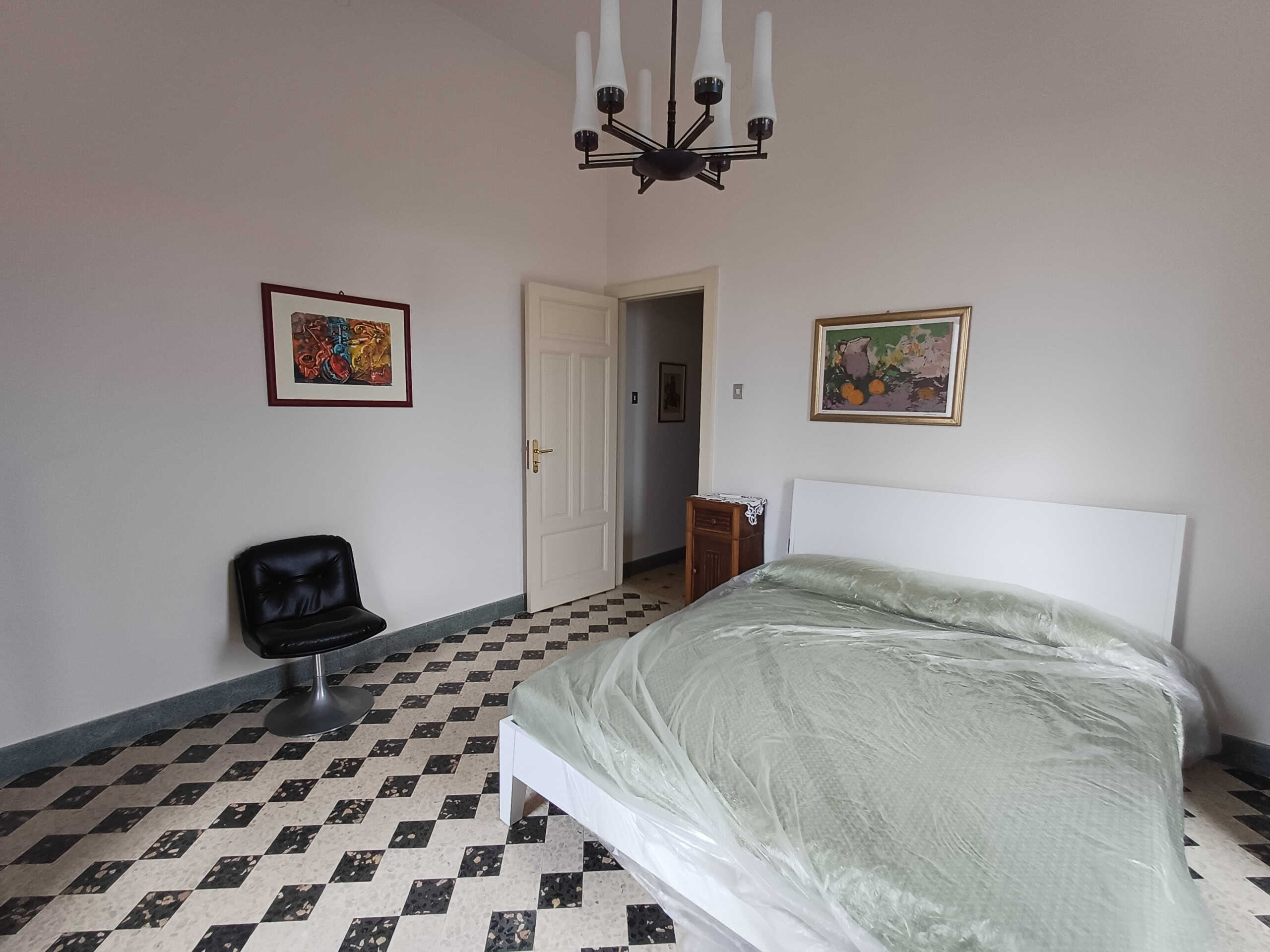 Camera da letto moderna con letto matrimoniale, pareti bianche, quadri decorativi, lampadario elegante, pavimento in piastrelle geometriche, arredamento funzionale, ambiente luminoso e confortevole, stile classico e raffinato, immagine di interni abitativi, soluzione immobiliare NextCasa.