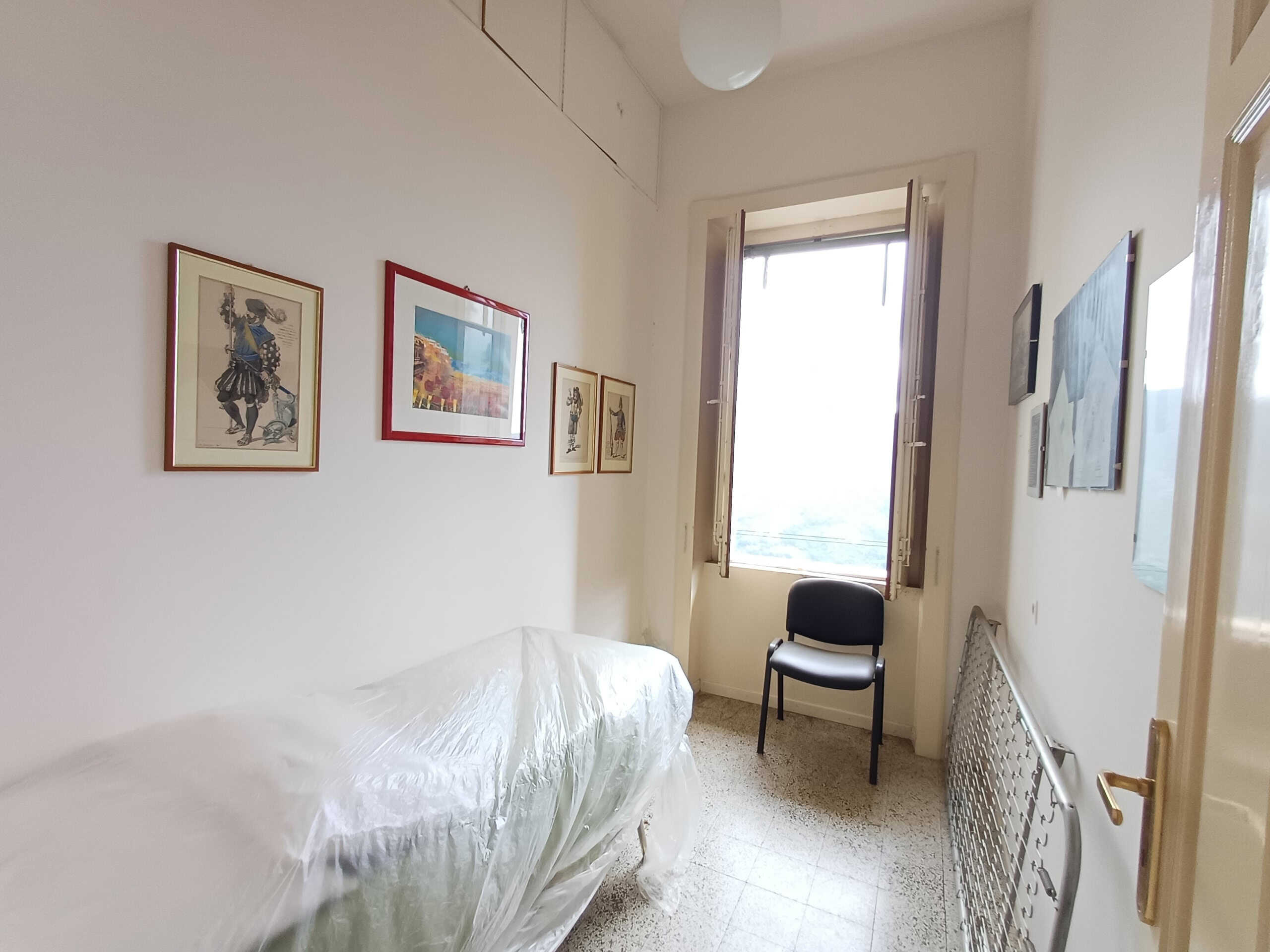 Soggiorno con pareti bianche, quadri decorativi e finestra con persiane, arredamento minimale con sedie e letto protetto, luce naturale, aria luminosa e spaziosa, ideale per soggiorni accoglienti.
