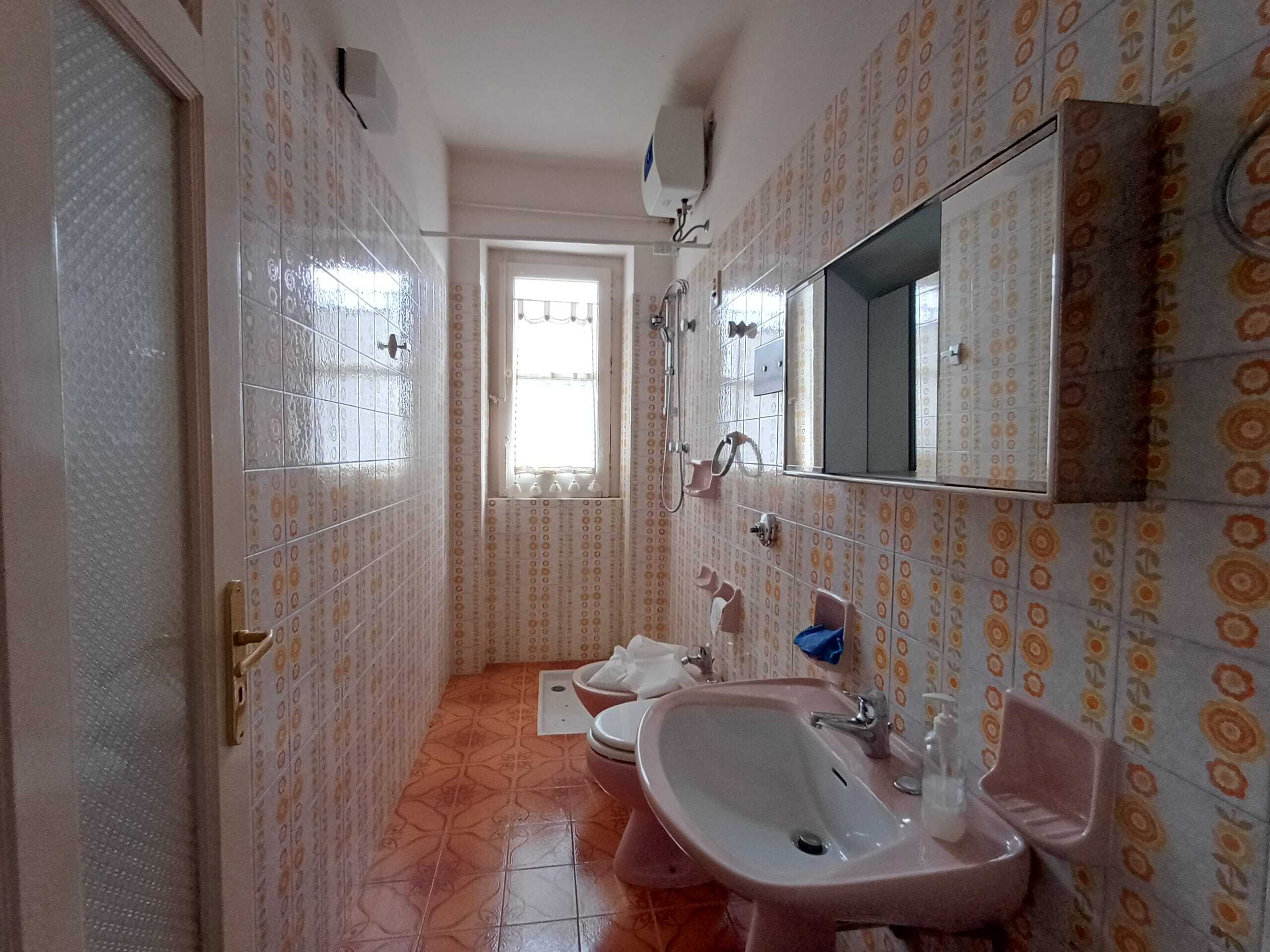 Lavandino da bagno rosa con specchio grande fronte, pareti e pavimento in piastrelle vintage con motivo arancione e giallo, finestra con tende leggere, elementi di arredo e accessori bagno.