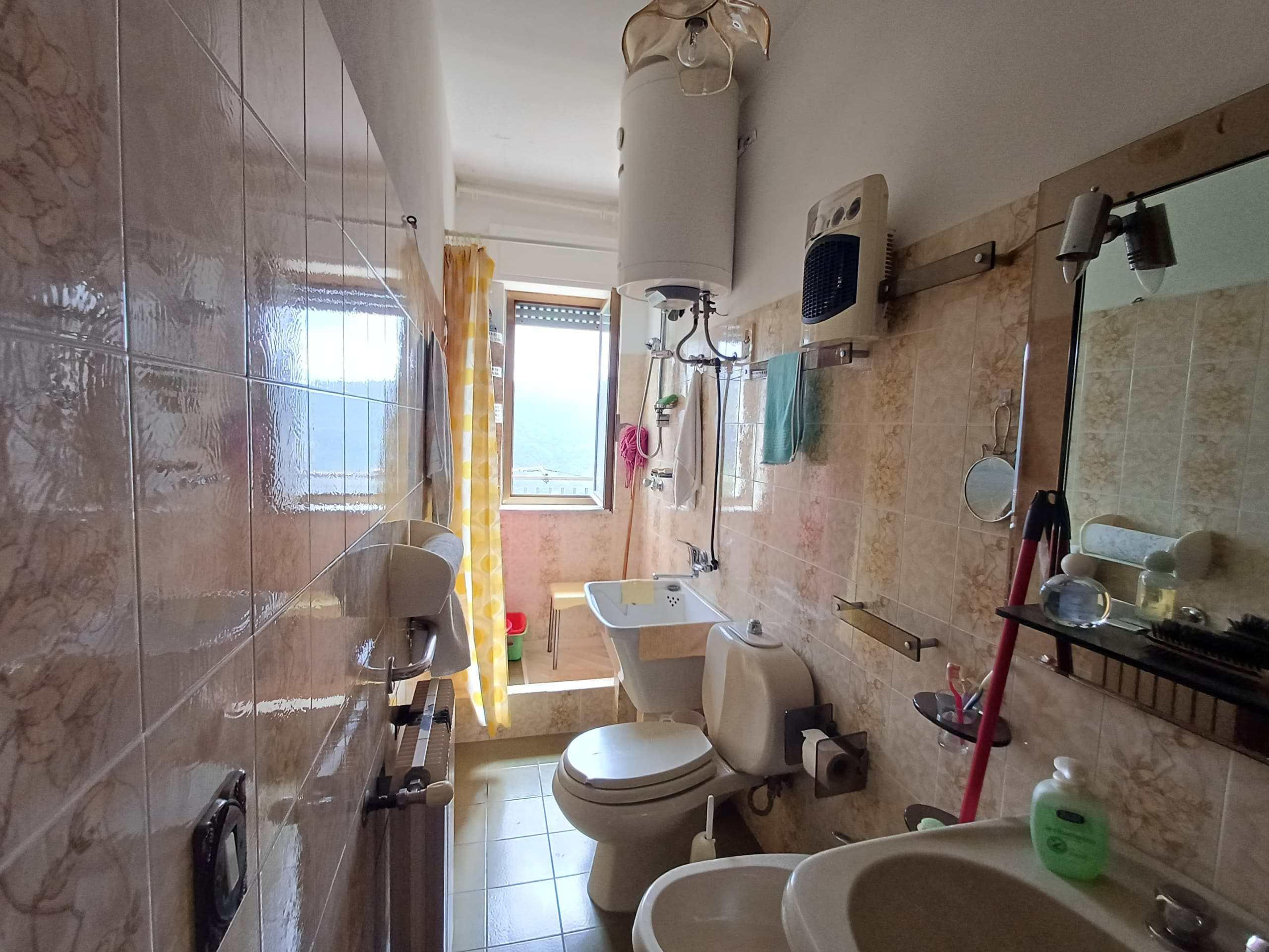 Lavabo, WC, e lavatrice in bagno con pareti e pavimenti in piastrelle beige, finestra con tenda gialla, numerosi accessori e oggetti di uso quotidiano.