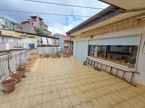 Balcone grande con piastrelle beige, ambiente esterno in stile urbano con vaso di fiori e ringhiera di sicurezza, vista sulle case e palazzi cittadini, ideale per relax e momenti all'aperto.