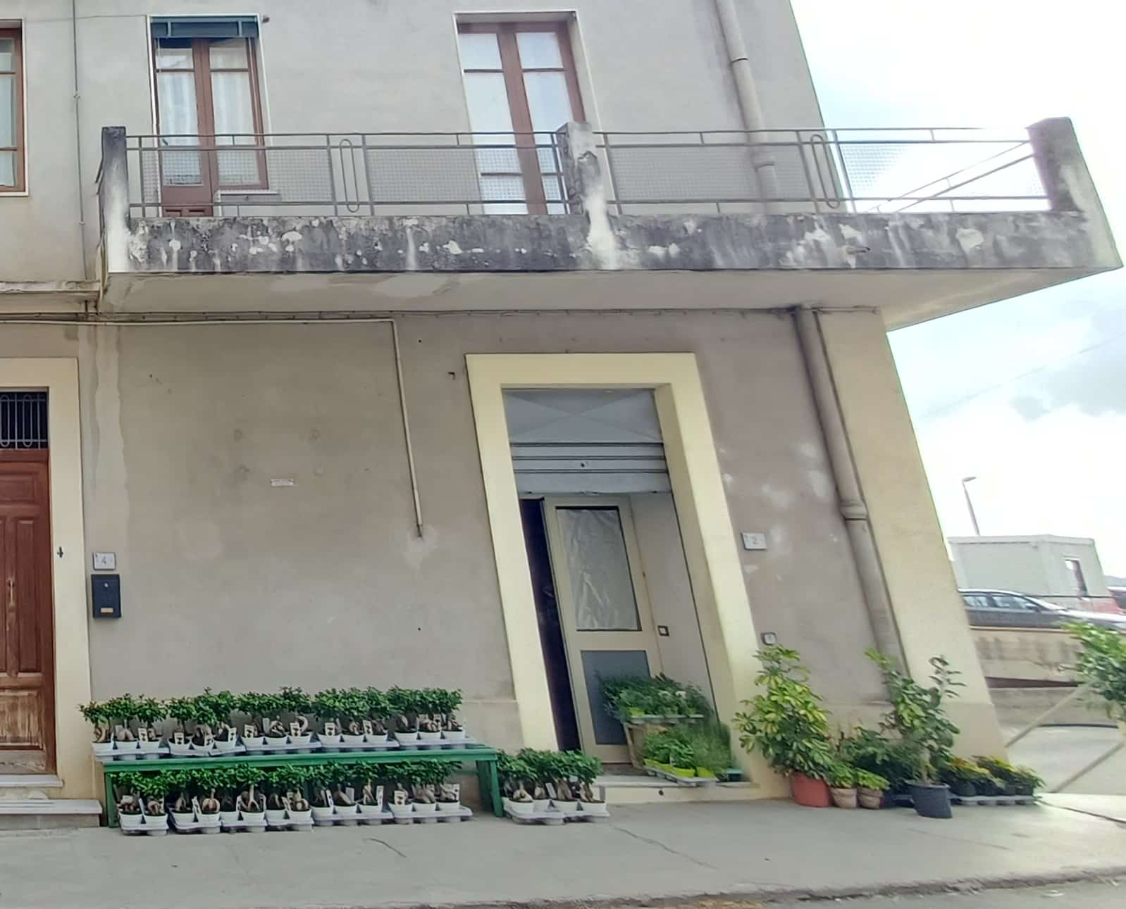 Ingresso di un appartamento con piante ornamentali su scaffale e ai lati della porta, facciata esterna in stile moderno, balcone con ringhiera in metallo, edificio con finiture in cemento e pareti color carne.