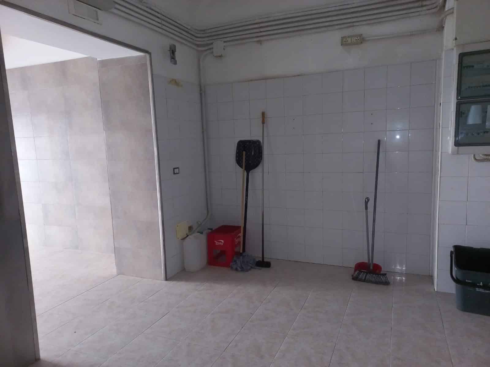 Spazio di lavoro o deposito con pareti rivestite in piastrelle bianche e pavimento in gres beige, attrezzato con scope, secchio e contenitori di plastica, adatto per servizi di pulizia e gestione ambienti.