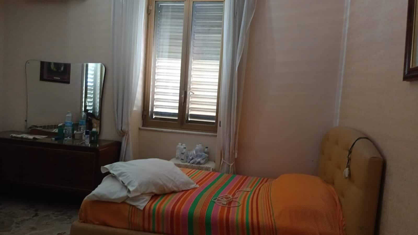 Letto singolo in soggiorno con finestra e mobili classici, ambiente accogliente e luminoso.