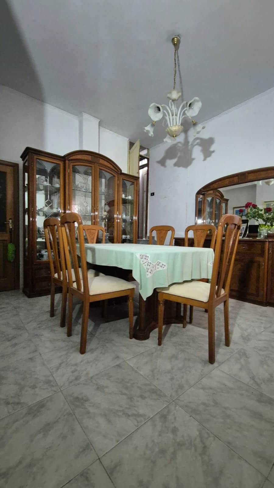Settimana da pranzo con tavolo e sedie in legno antico, vetrinetta in legno con cristalli, lampadario classico, arredamento tradizionale, ambiente interno caldo e accogliente, stile casa di famiglia.