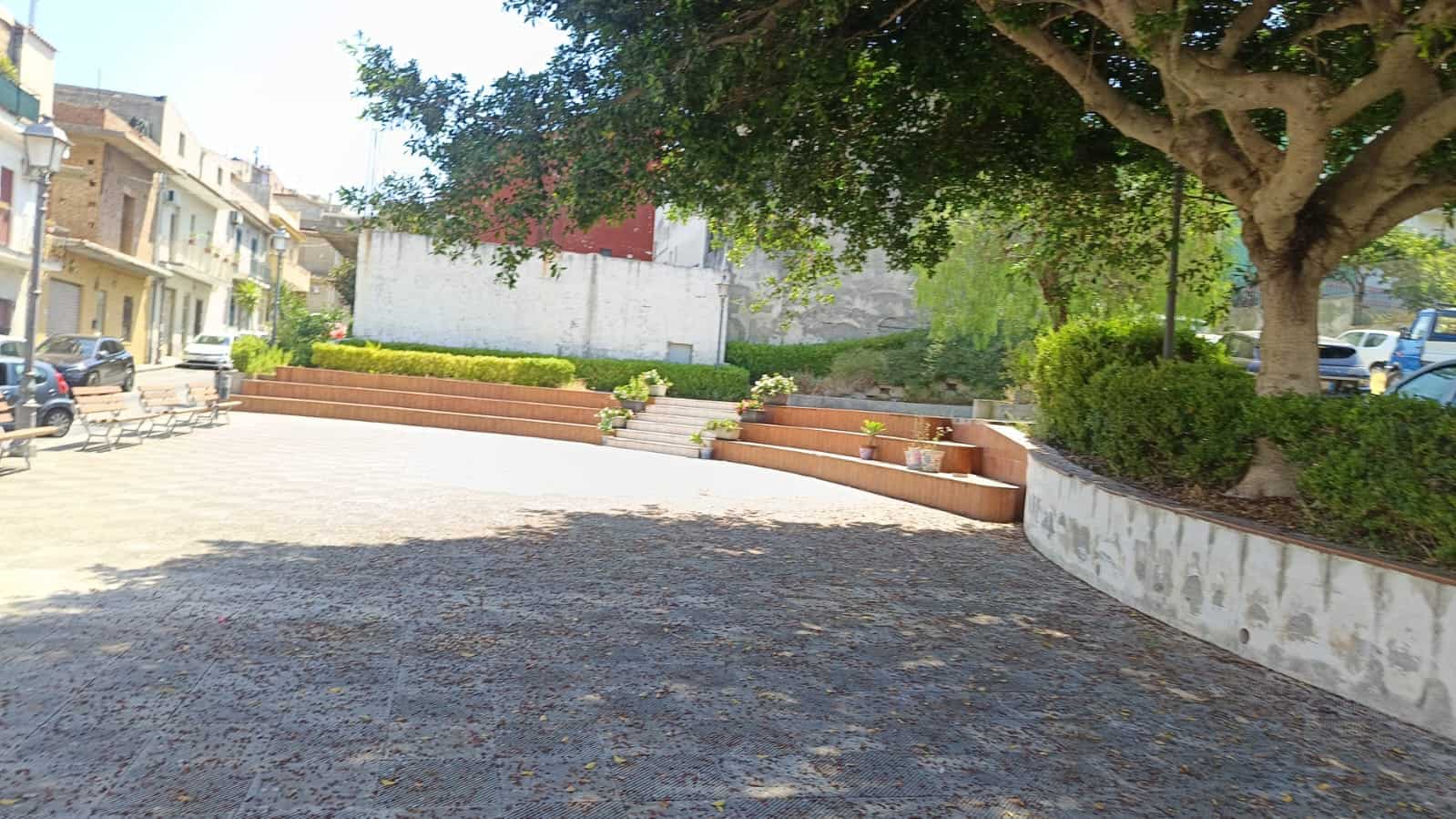 Area esterna di piazzale con panchine e piante, spazio pubblico ben curato e ombreggiato da alberi, ideale per relax e incontri all'aperto vicino alle abitazioni.