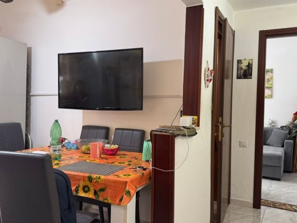 L'immagine mostra una zona pranzo in un appartamento con un tavolo ricoperto da una tovaglia arancione con girasoli, sedie nere imbottite, e una parete con televisore a schermo piatto. Si notano anche una cucina con frigorifero decorato con magneti e una zona con porta che conduce ad un soggiorno. L'ambiente è accogliente e funzionale, ideale per convivialità domestica.