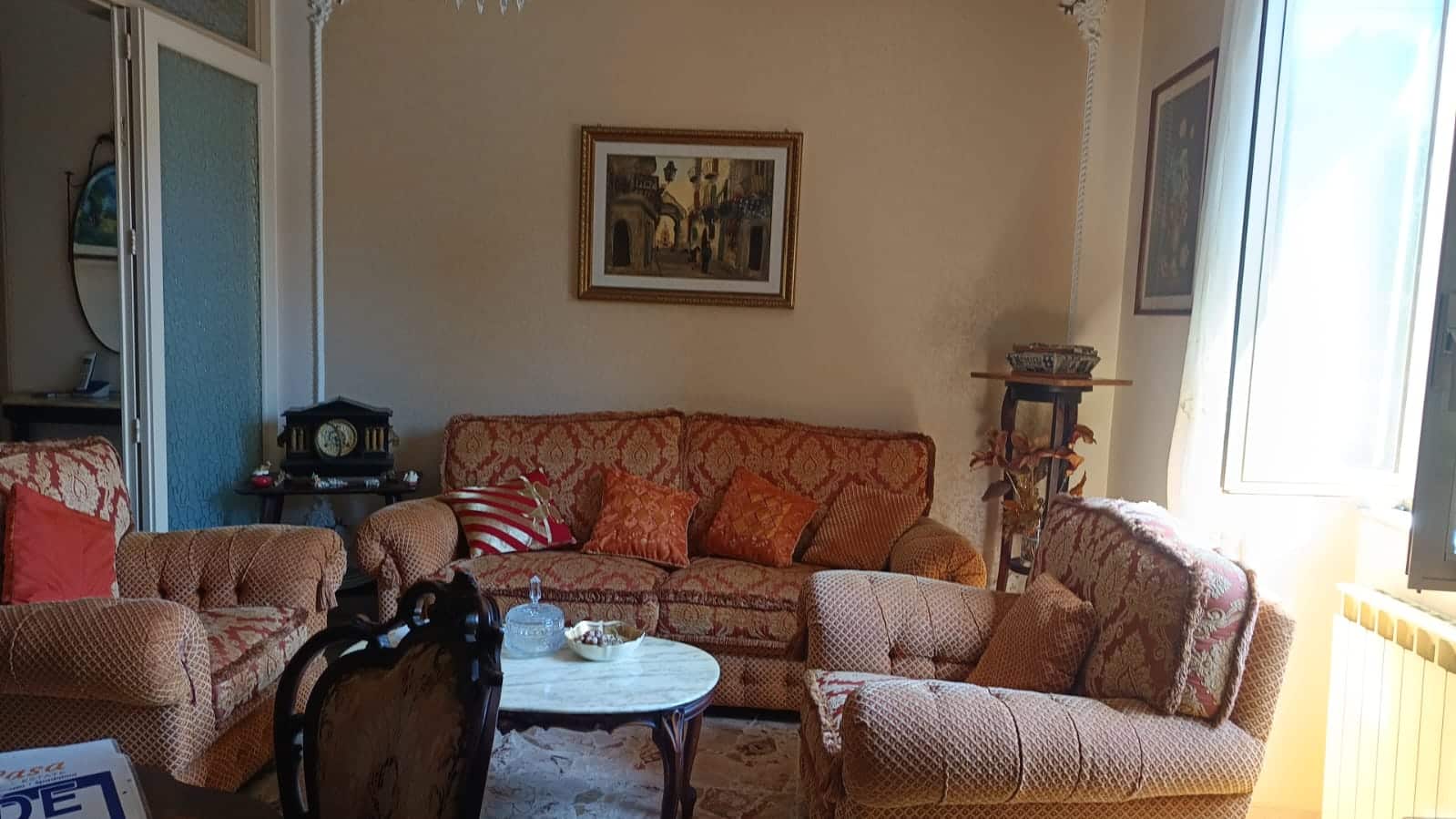 Soggiorno elegante con divani imbottiti rivestiti in tessuto damascato, decorazioni classiche, cornice dorata e affreschi d'epoca, luce naturale da finestra, atmosfera accogliente e raffinata.