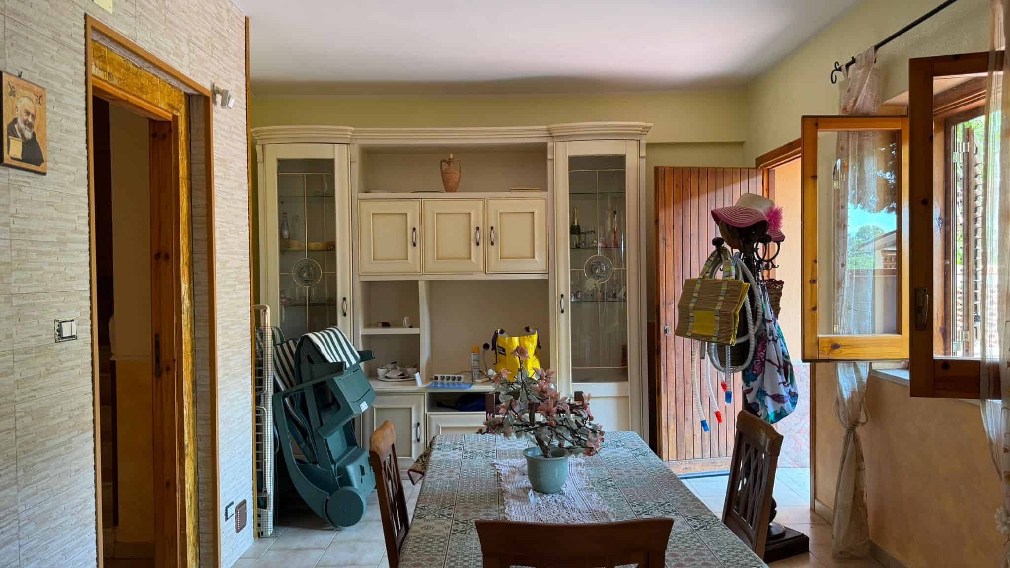 Armadio in stile classico con vetrinetta e tavolo da pranzo in cucina, ambiente accogliente con decorazioni, finestre aperte e luce naturale, arredamento caldo e funzionale, casa familiare.