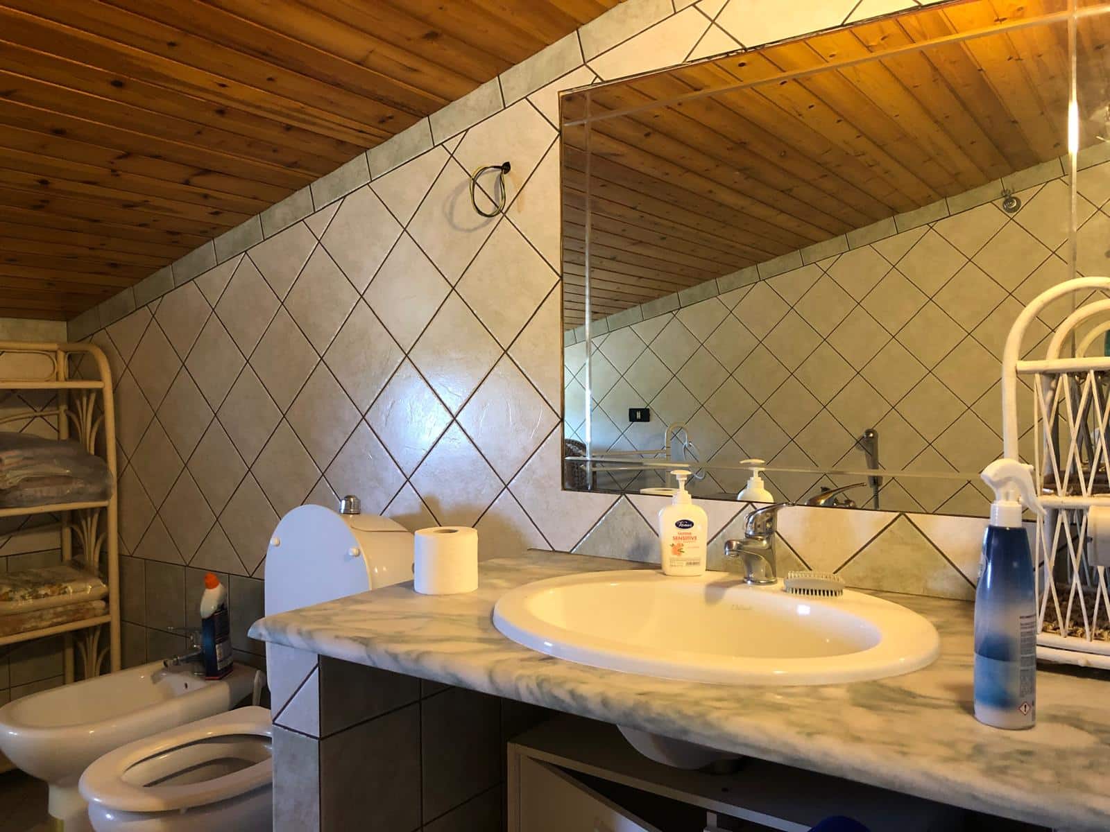 Lavabo in bagno con specchio grande, rumenti e prodotti per la pulizia, soffitto in legno e pareti rivestite in piastrelle beige, ambiente accogliente e funzionale.