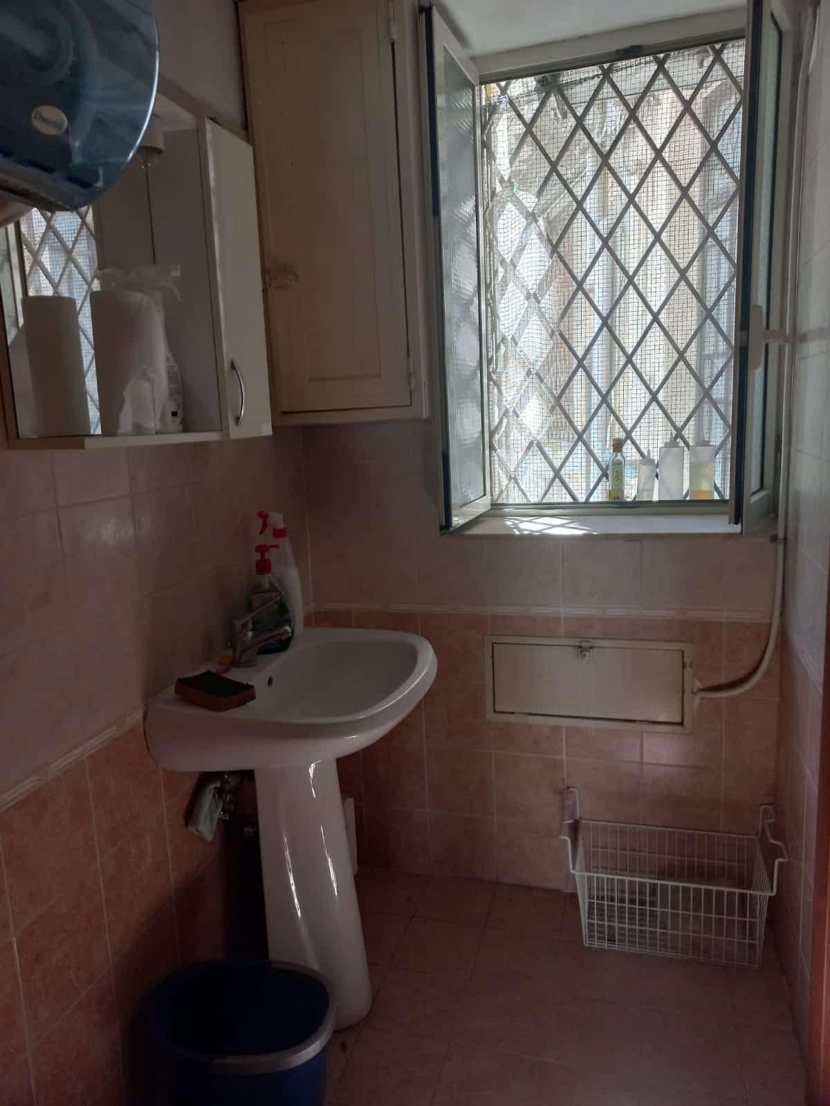 Lavabo bianco in bagno con finestra e sbarre di sicurezza, arredi essenziali, crema e beige toni, accessori per la pulizia e spazio di stoccaggio, atmosfera semplice e funzionale.