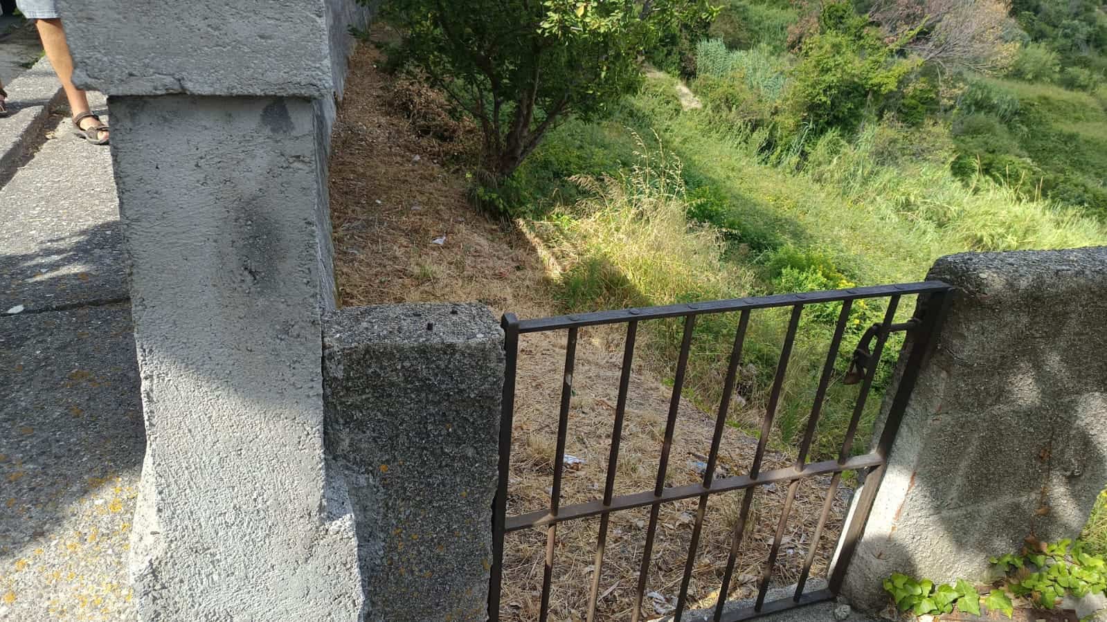 Fioriera in pietra e recinzione in ferro su un tratto di strada di campagna con vista su un paesaggio verde e collinare.