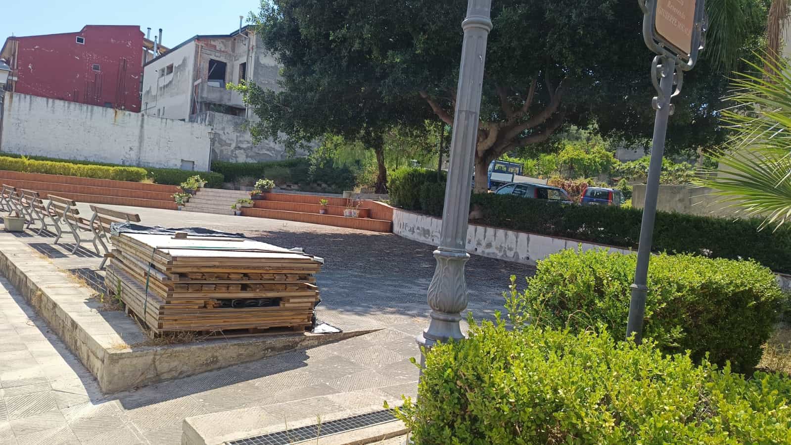 Panchina e piante nel parco pubblico con alberi e sedie in pietra, ambiente sereno e verdeggiante, adatto a rilassarsi all'aperto, presenti macchinari per lavori di costruzione e strada pavimentata.